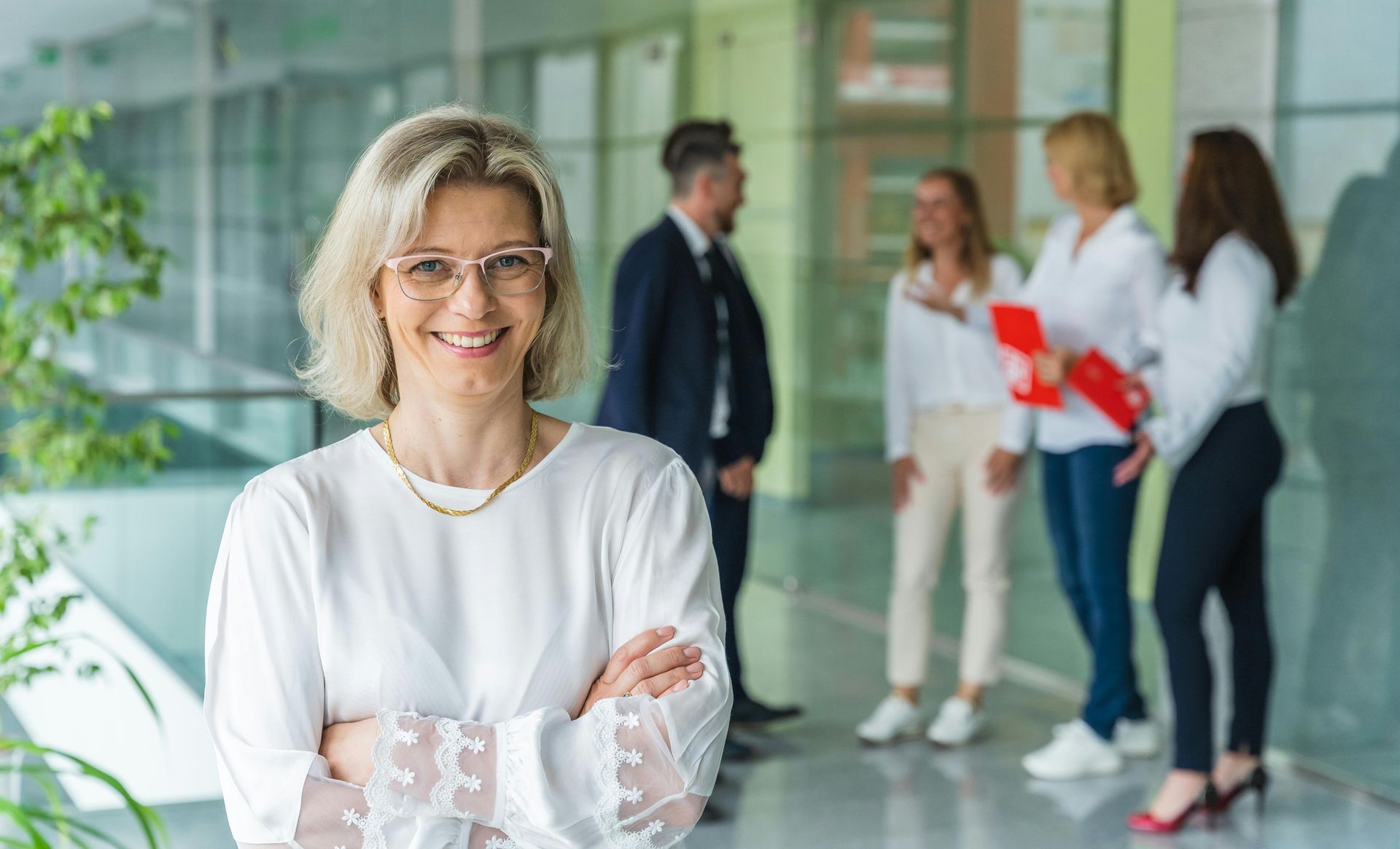 Woman in white blouse with arms crossed smiles confidently in an office setting. Group of people in background.