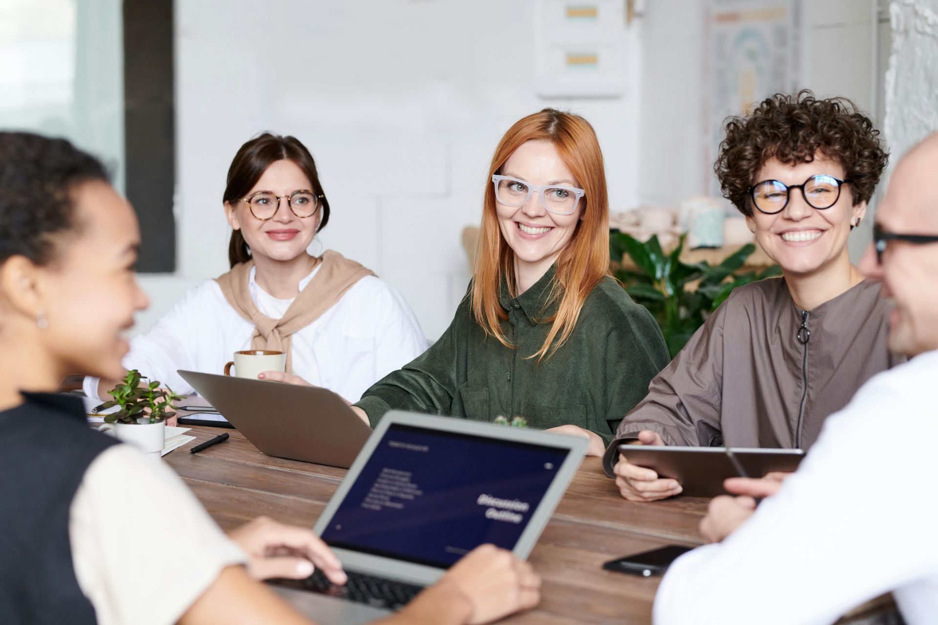 People smiling and working together at a wooden table with laptops and tablets.