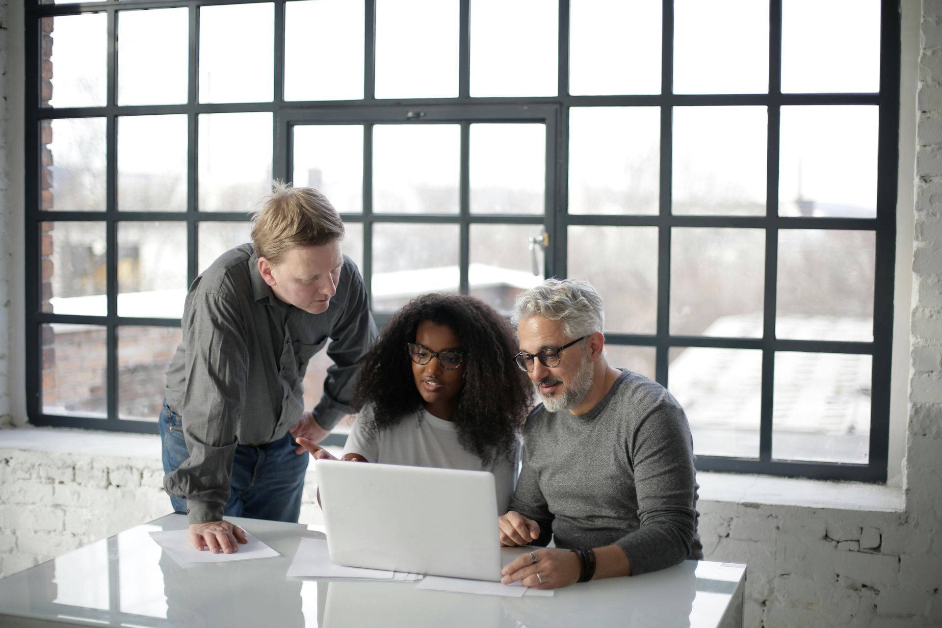 Three people looking at laptop screen in front of a window. One is leaning, others are seated.