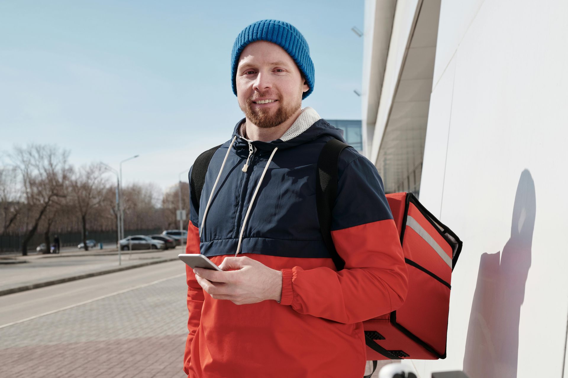 A food delivery person with a red insulated backpack standing outside, looking at a smartphone.