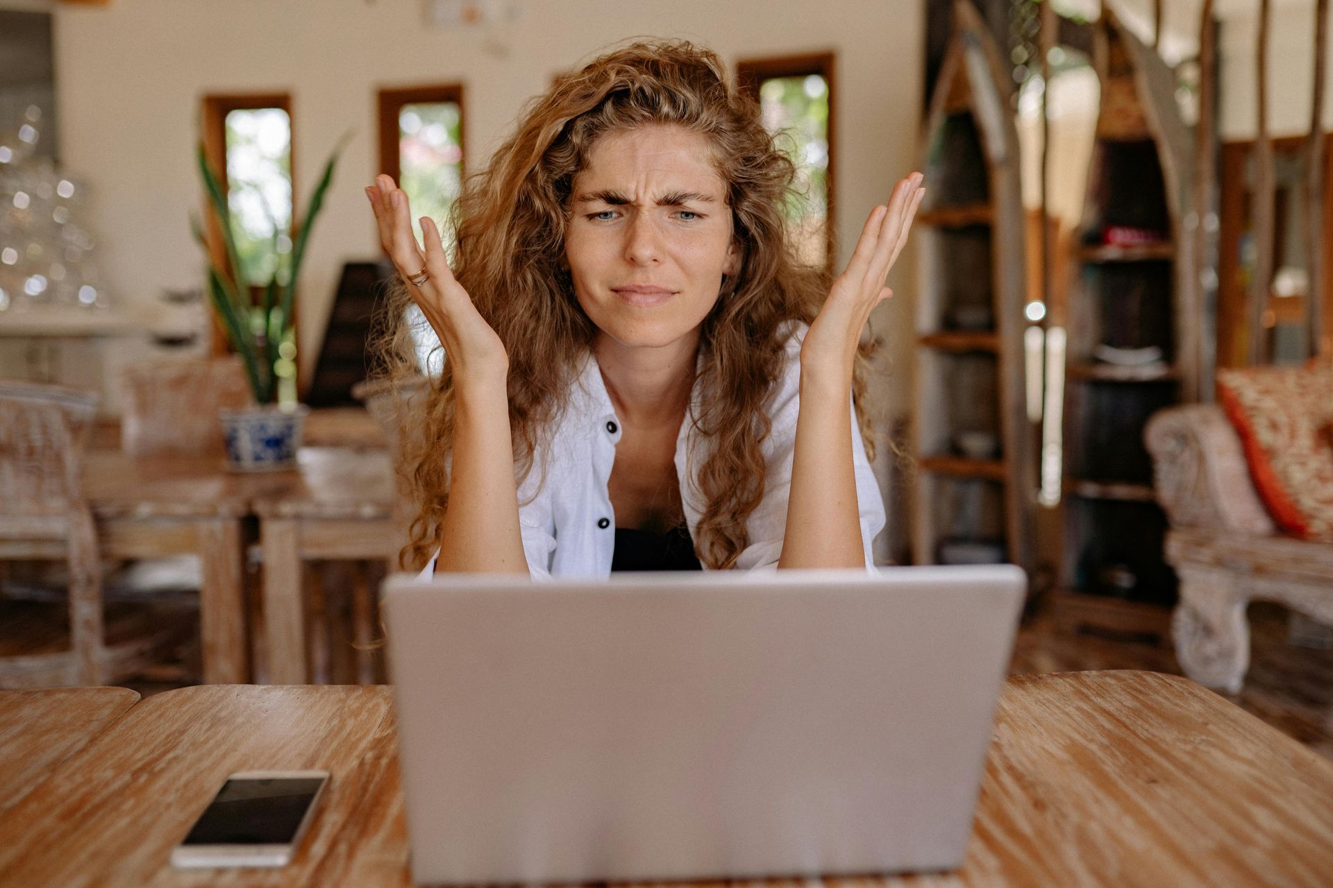 Woman with curly hair, hands raised in frustration, looking at a laptop on a wooden table.