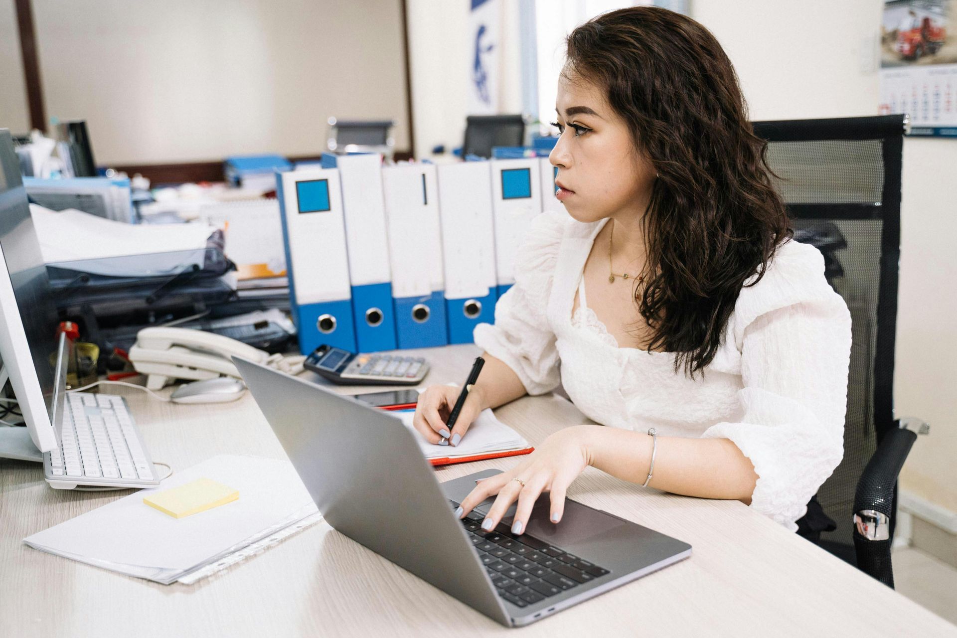 Woman at desk in office, working on laptop and writing in notepad.