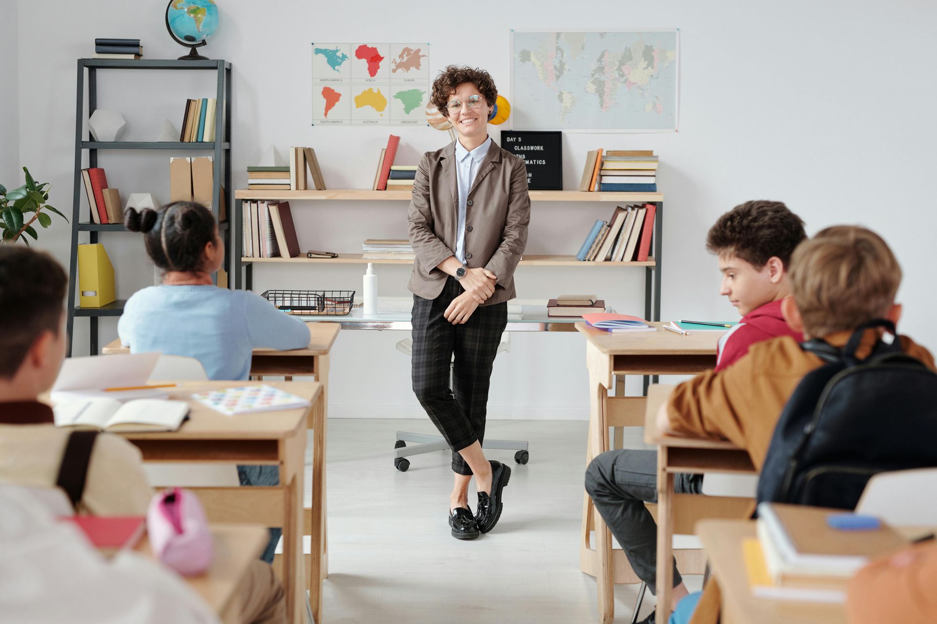 Teacher standing in a classroom smiles at students seated at desks. Bookshelves and world map background.