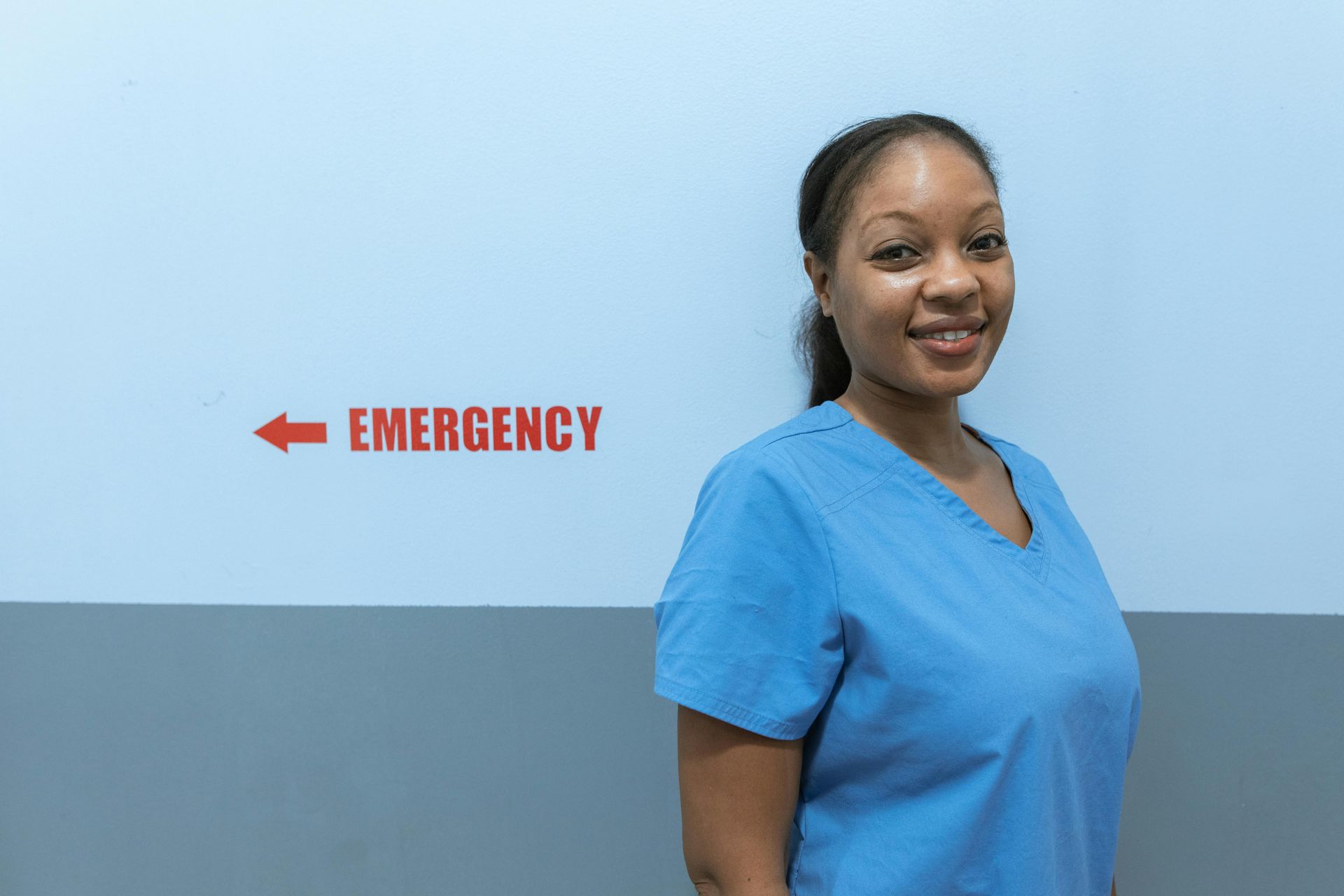 Woman in blue scrubs smiles in front of a wall with an emergency sign.