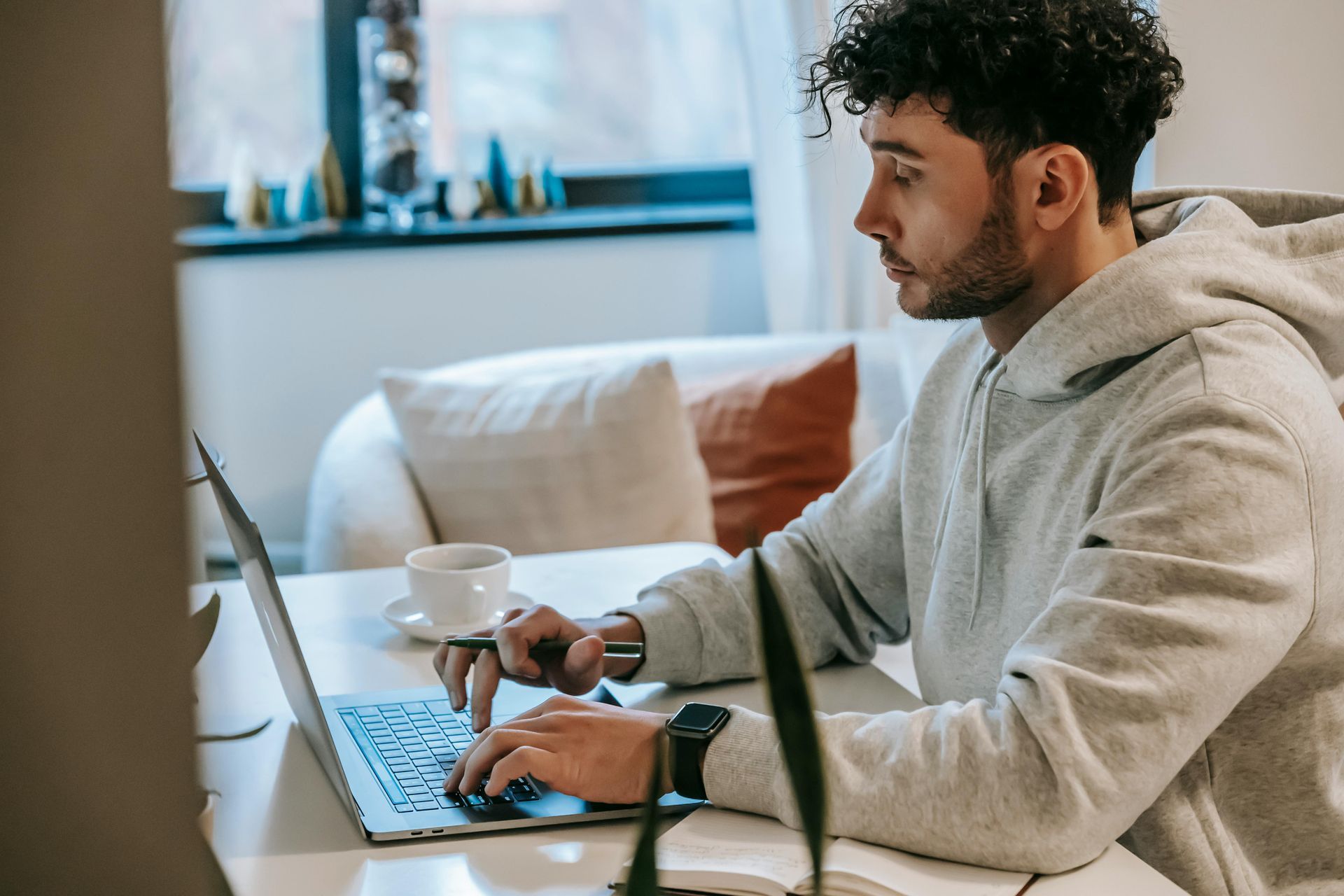 Man in a gray hoodie types on a laptop at a desk indoors. A mug and pen are nearby.