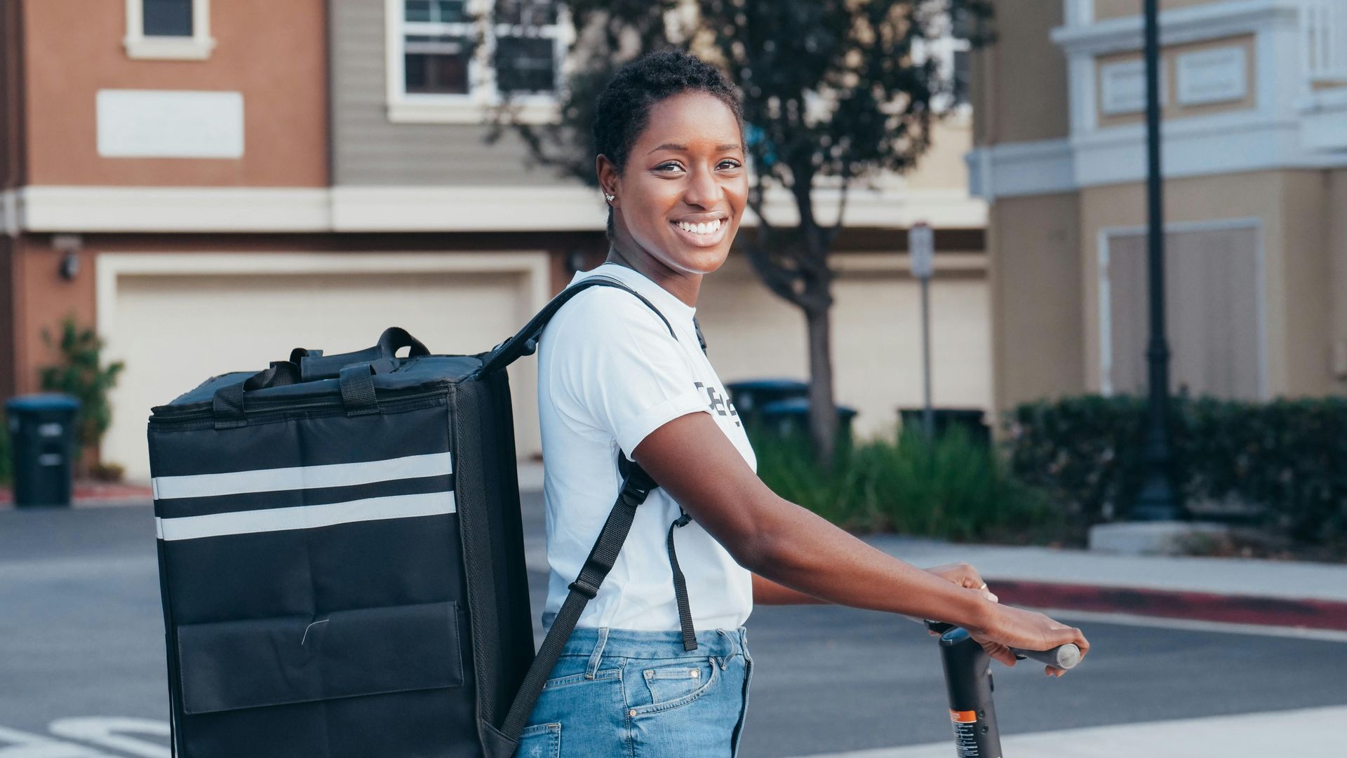 Woman smiling, riding a scooter with a delivery backpack, on a suburban street.
