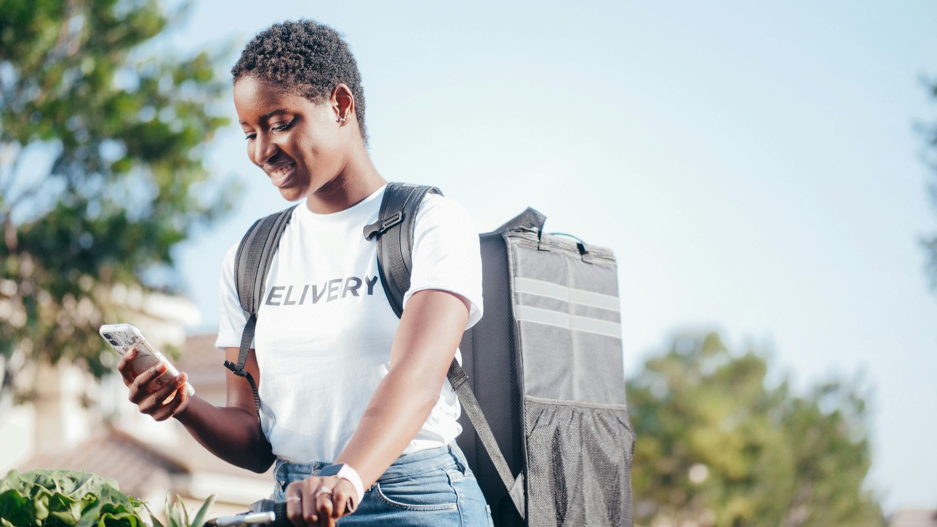 Woman with backpack, looking at phone, delivering food on a sunny day.