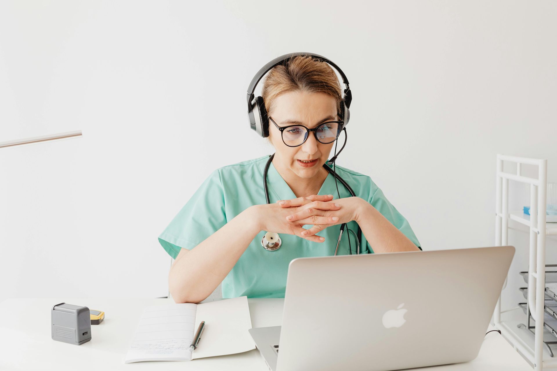 Medical professional in scrubs and glasses on a video call, wearing a headset, looking at a laptop.