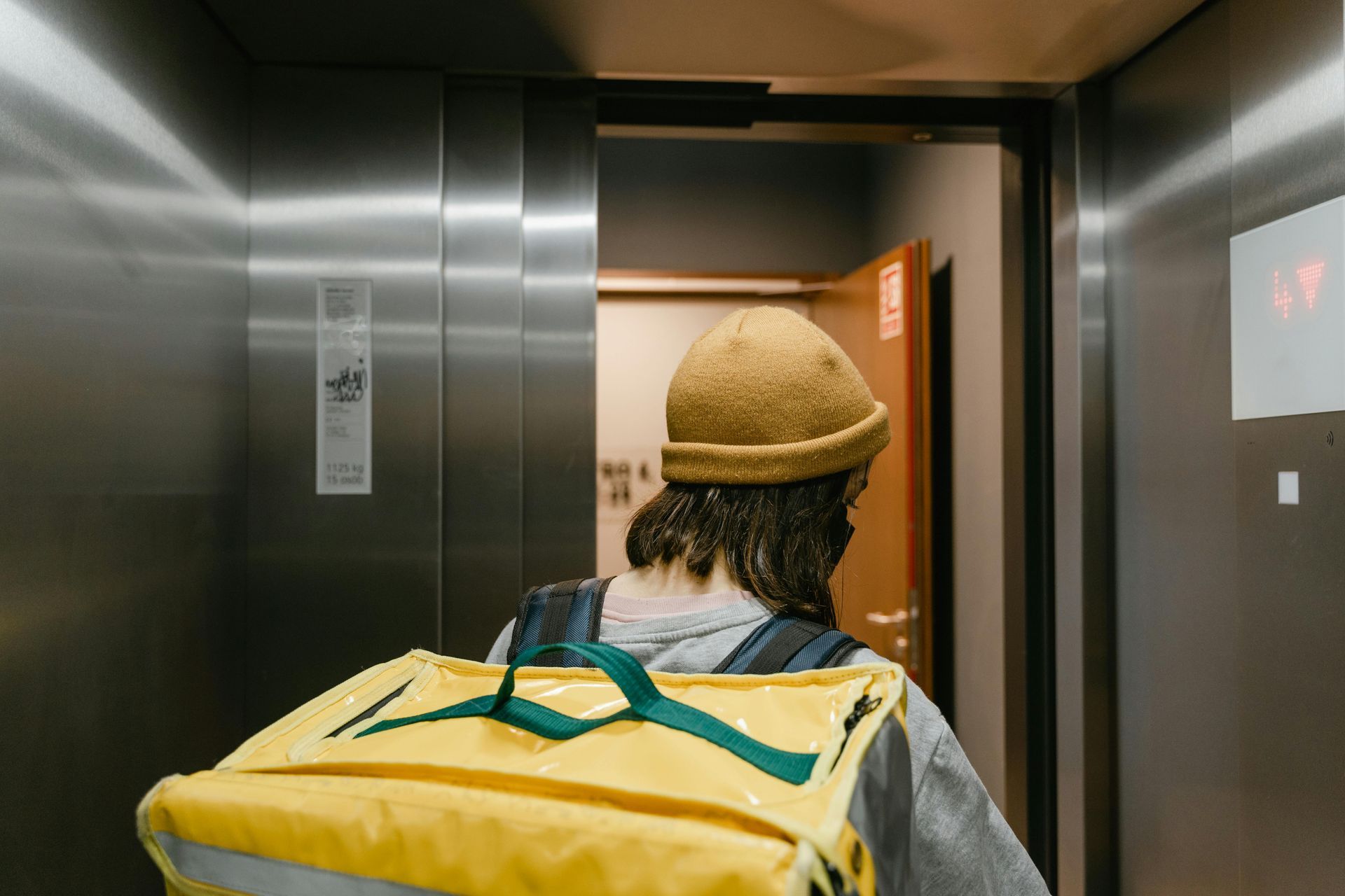 Person with a yellow delivery backpack exits an elevator into a hallway with a wooden door.