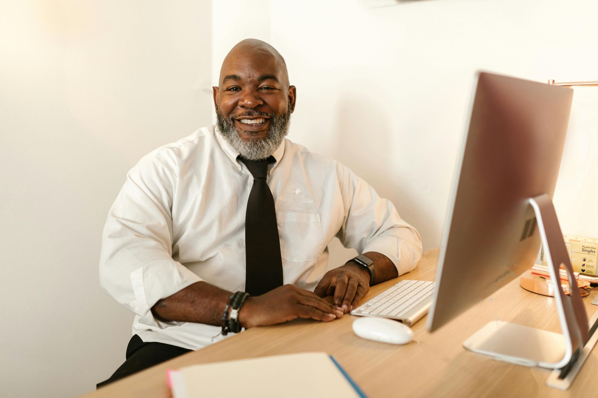 A smiling Black man with a gray beard sits at a desk with a computer.