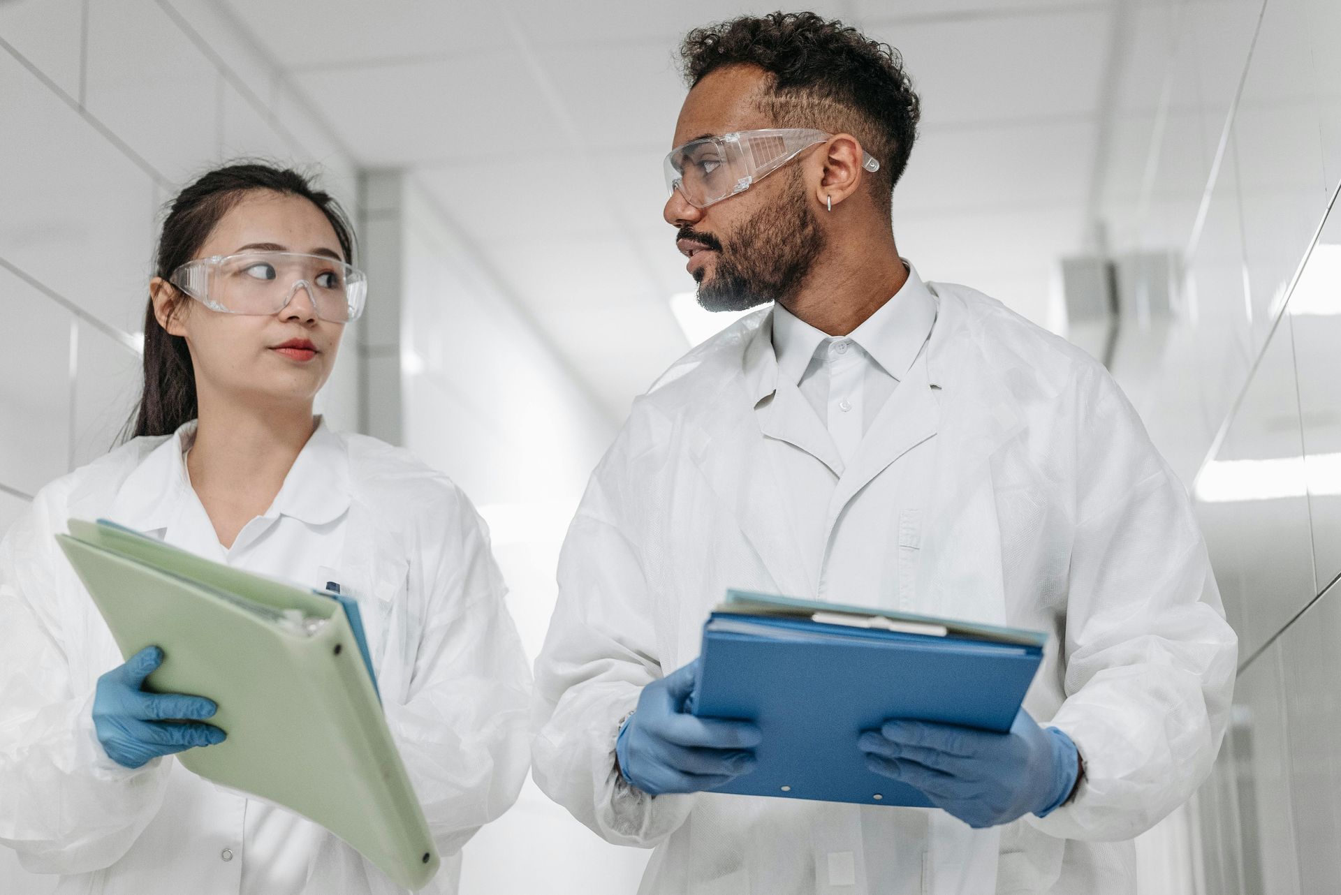 Two people in lab coats and safety glasses walk in a hallway, holding clipboards.