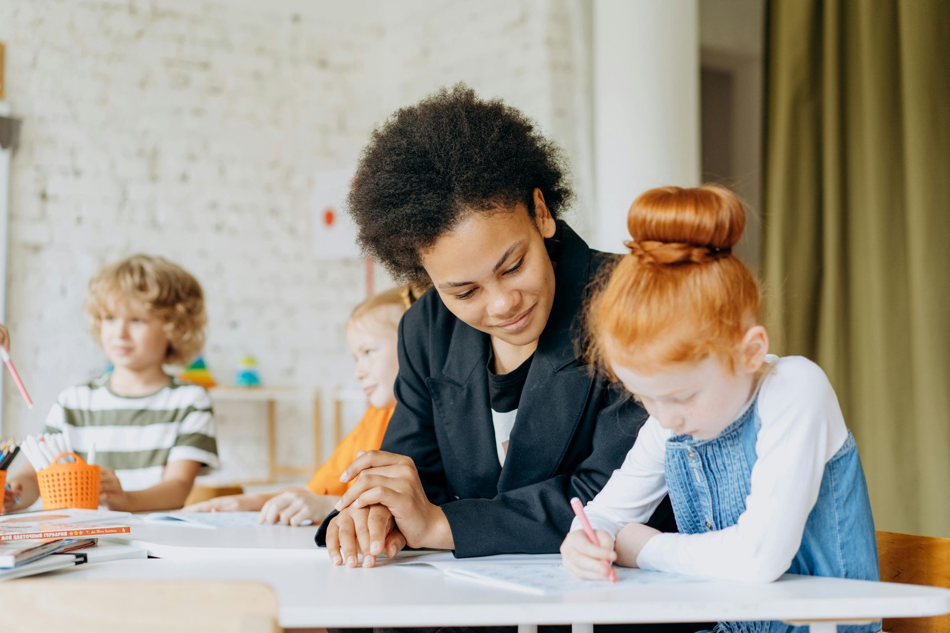 A teacher assists a young student with schoolwork at a classroom desk, while other children work in the background.