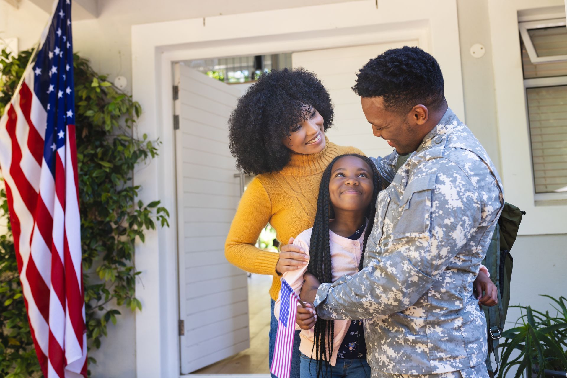 Soldier kneels to greet a person holding a laughing toddler in a living room.