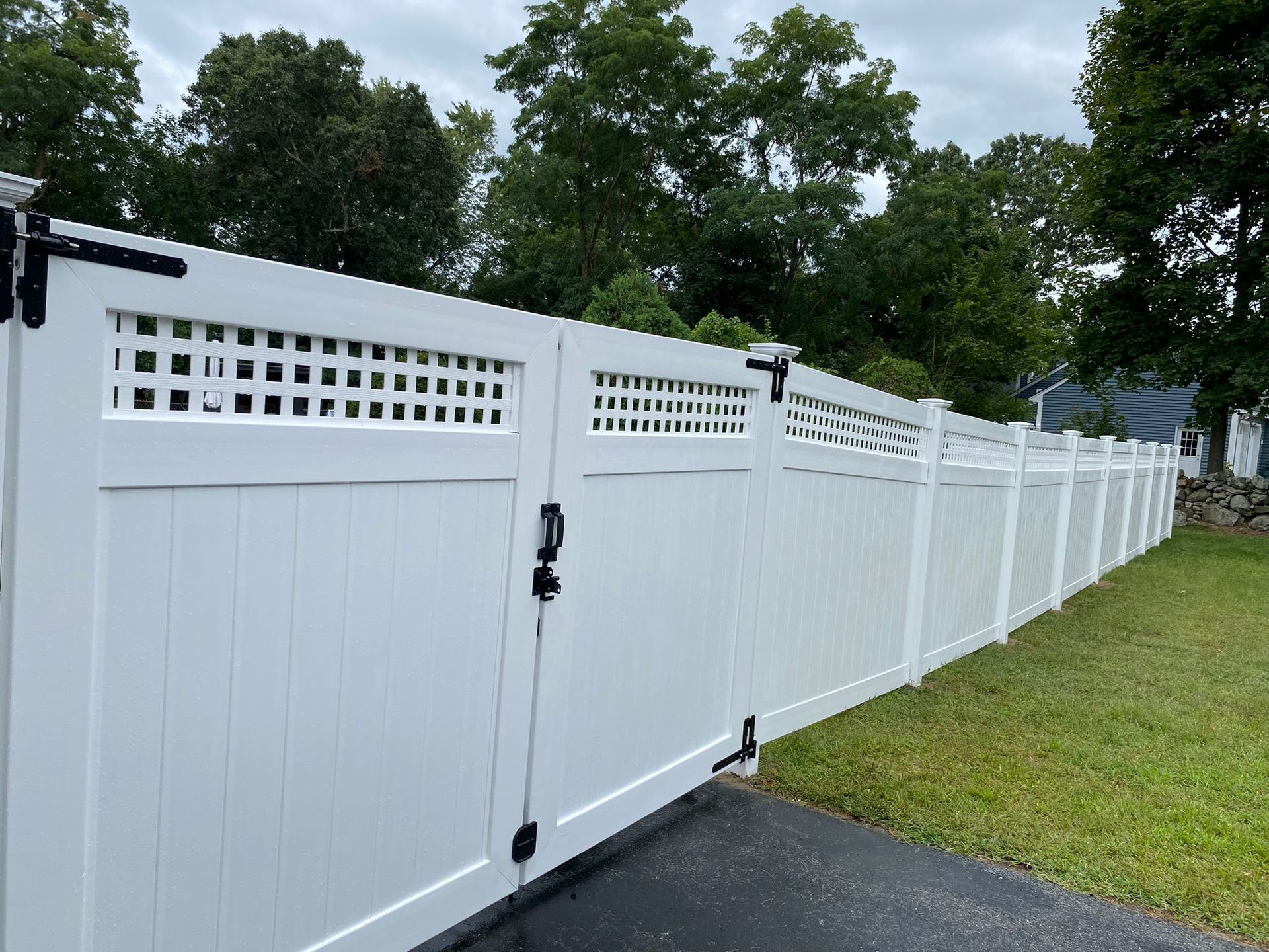 A white vinyl fence with a black gate is surrounded by grass and trees.