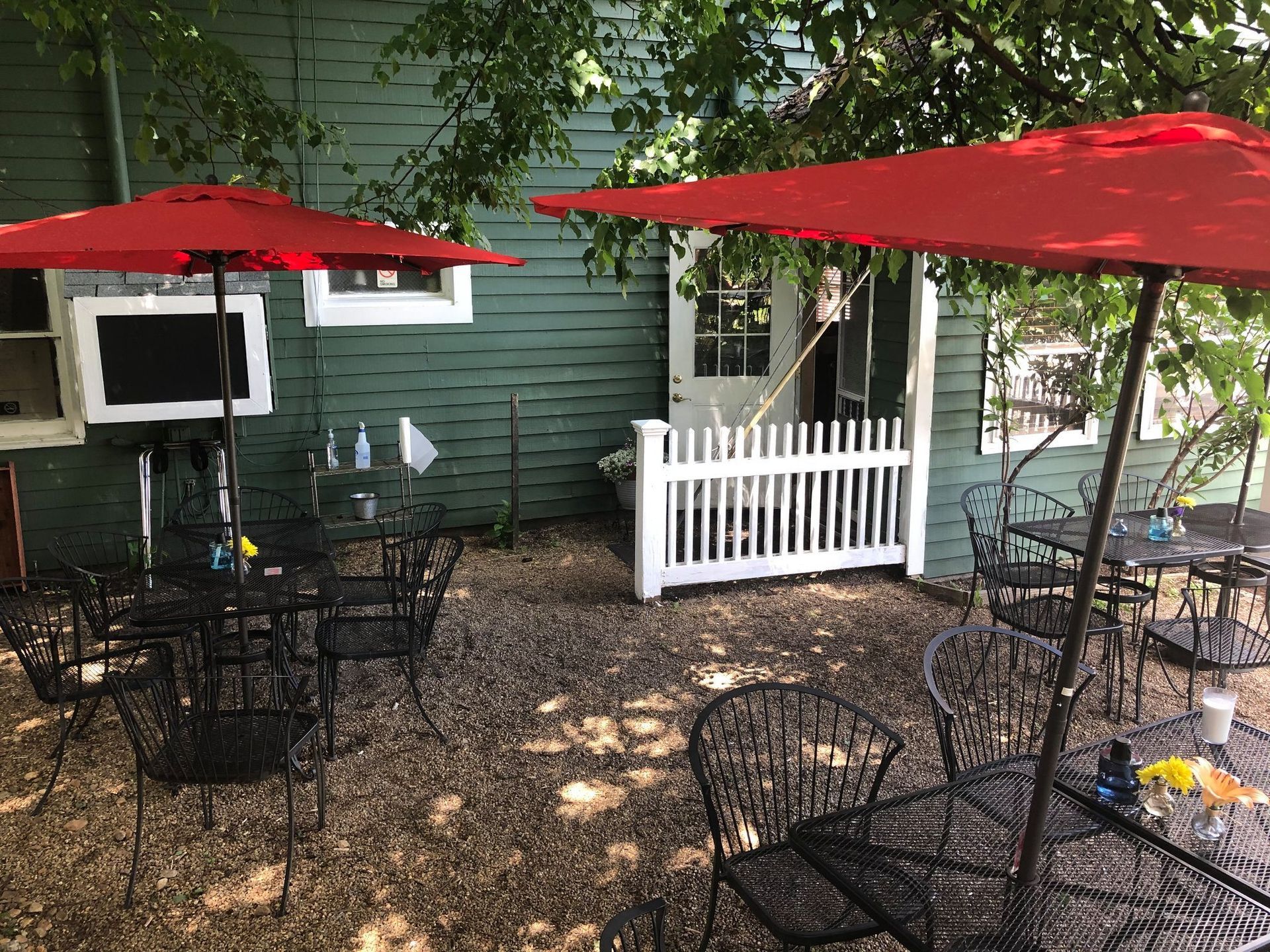 Outdoor dining area with tables and black metal chairs under red umbrellas next to a green building with a white fence.