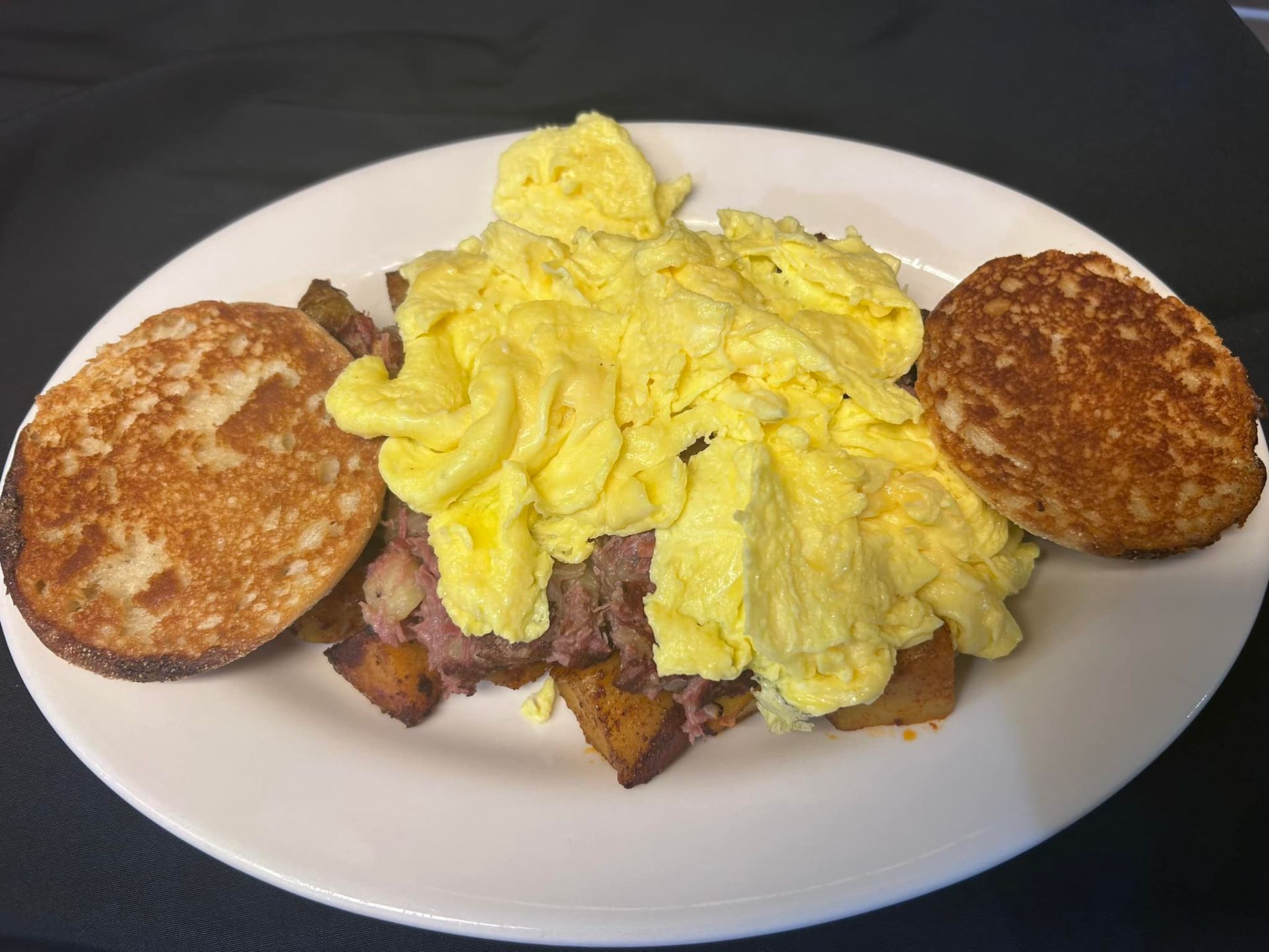 A plate of breakfast food featuring scrambled eggs atop corned beef hash, flanked by two halves of a toasted English muffin.