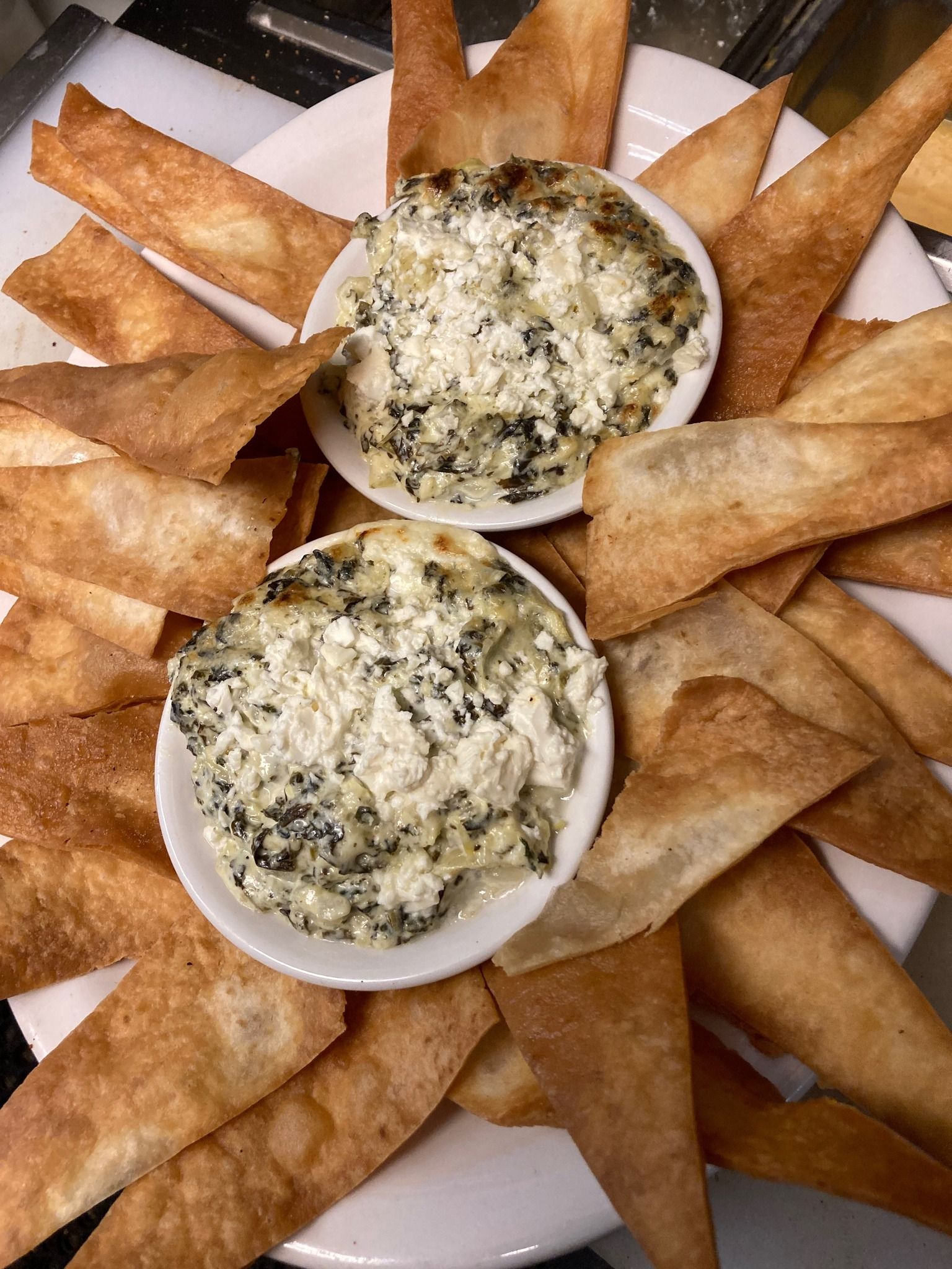 Two white bowls of spinach artichoke dip surrounded by crispy, golden-brown triangular tortilla chips on a white plate.