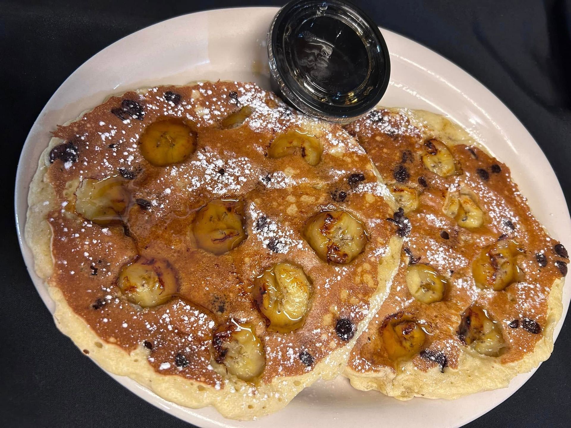 Two pancakes topped with sliced bananas, chocolate chips, and powdered sugar, served on a plate with a side of syrup.