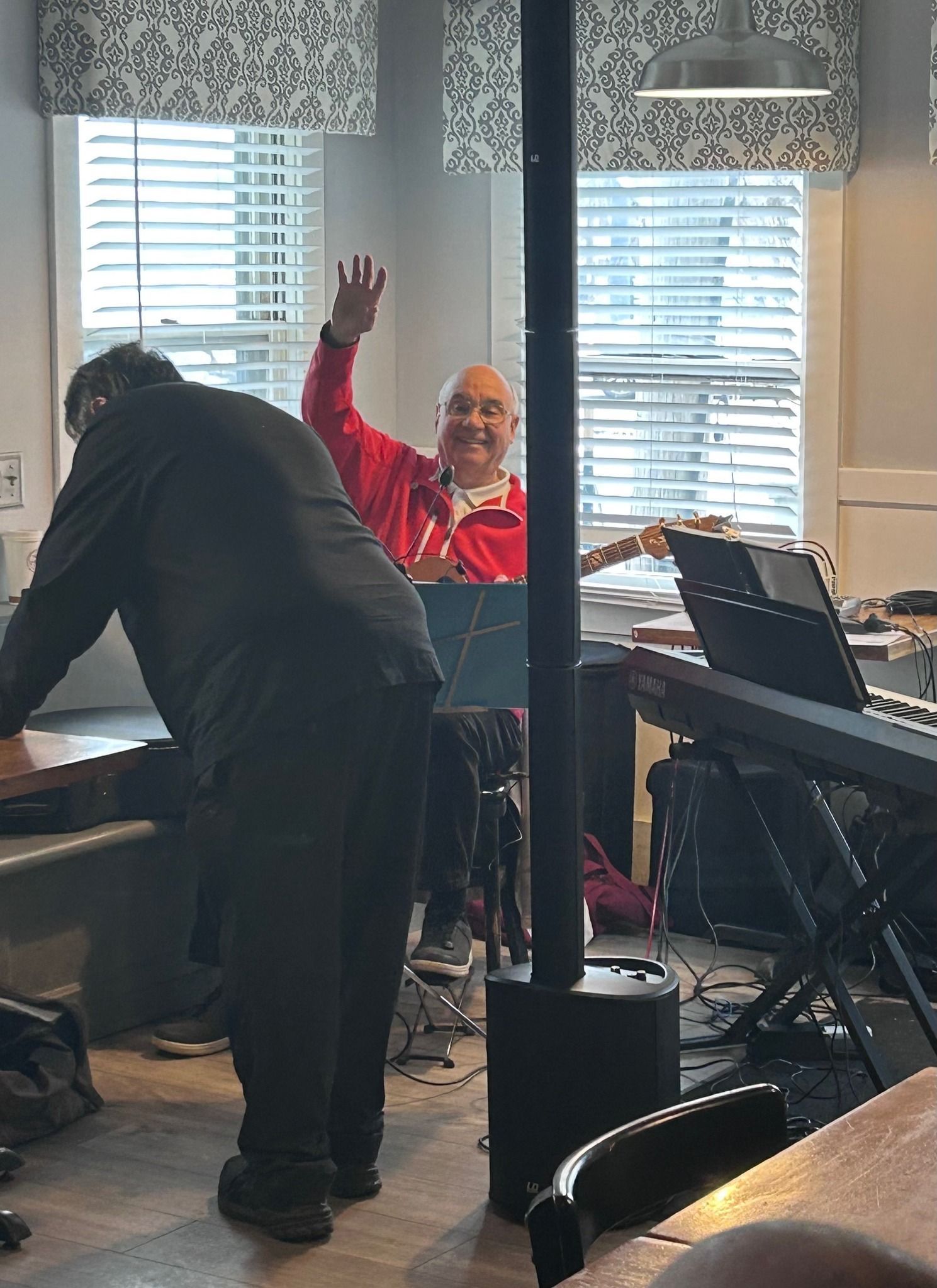 A person in a black outfit stands at a piano while another in a red shirt plays guitar and waves in a room with blinds.