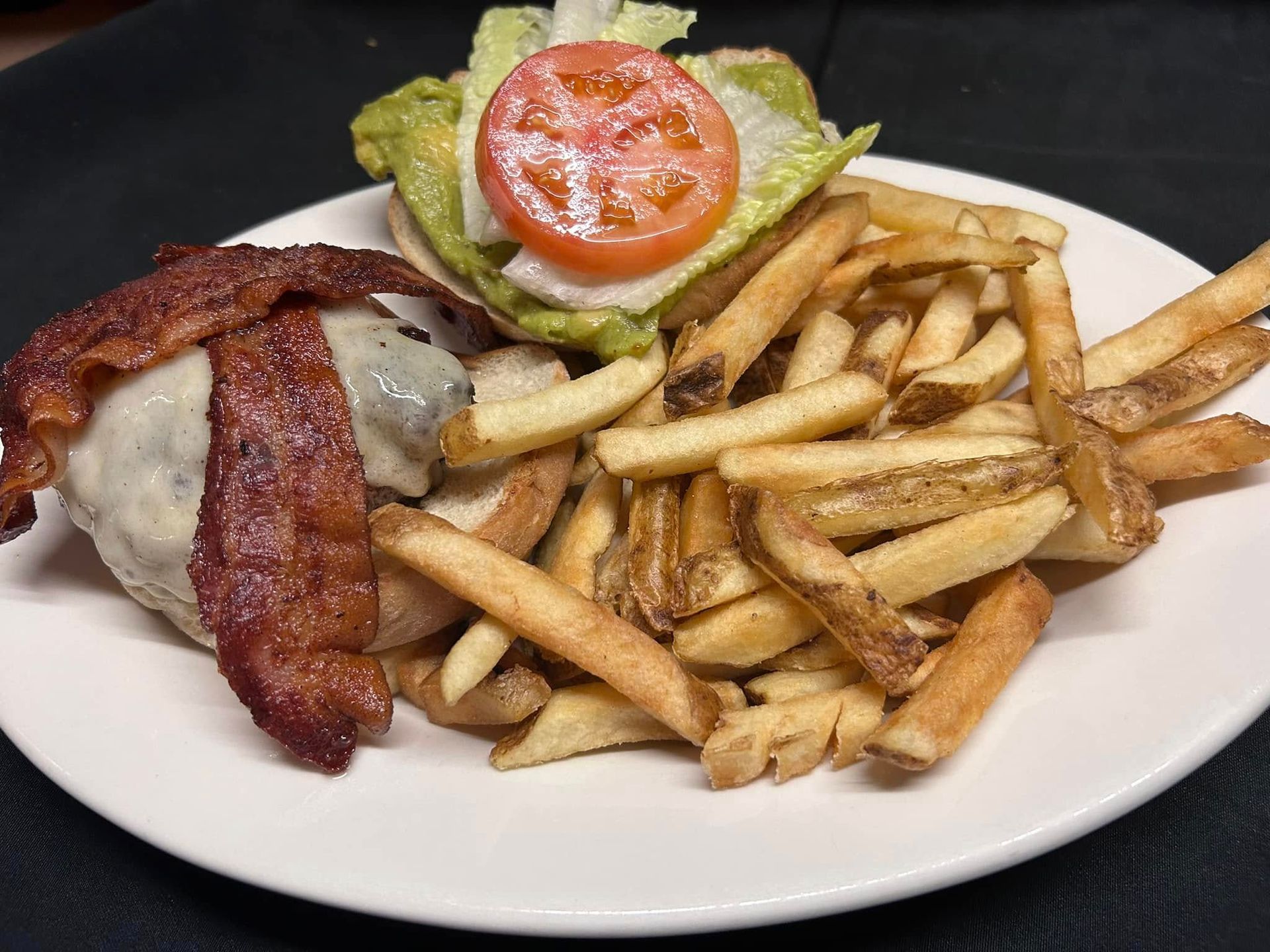 A bacon cheeseburger with a tomato slice and avocado on top, served with a side of french fries on a white plate.