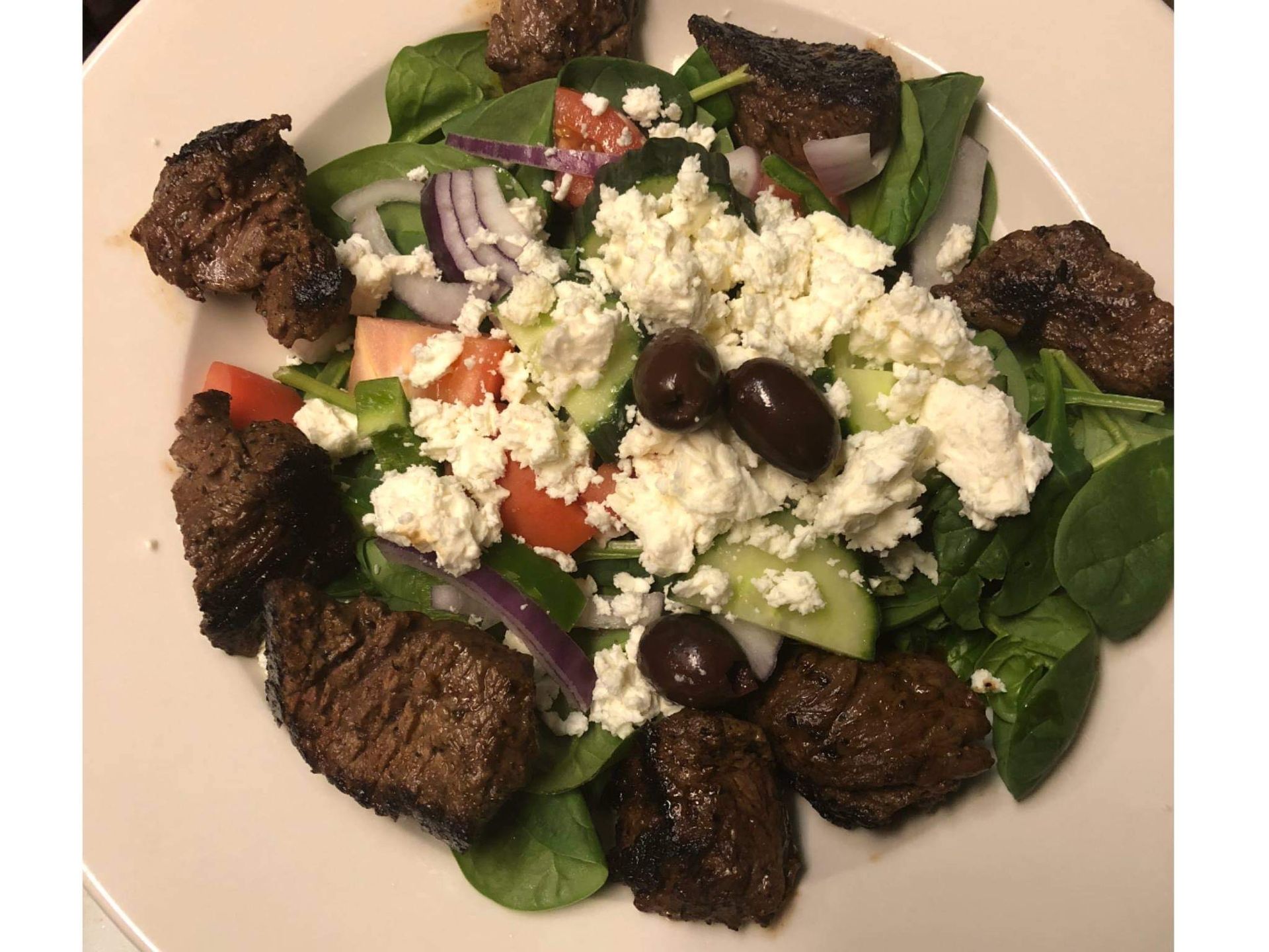 A steak salad with spinach, feta cheese, olives, red onion, tomatoes, and cucumber on a white plate.