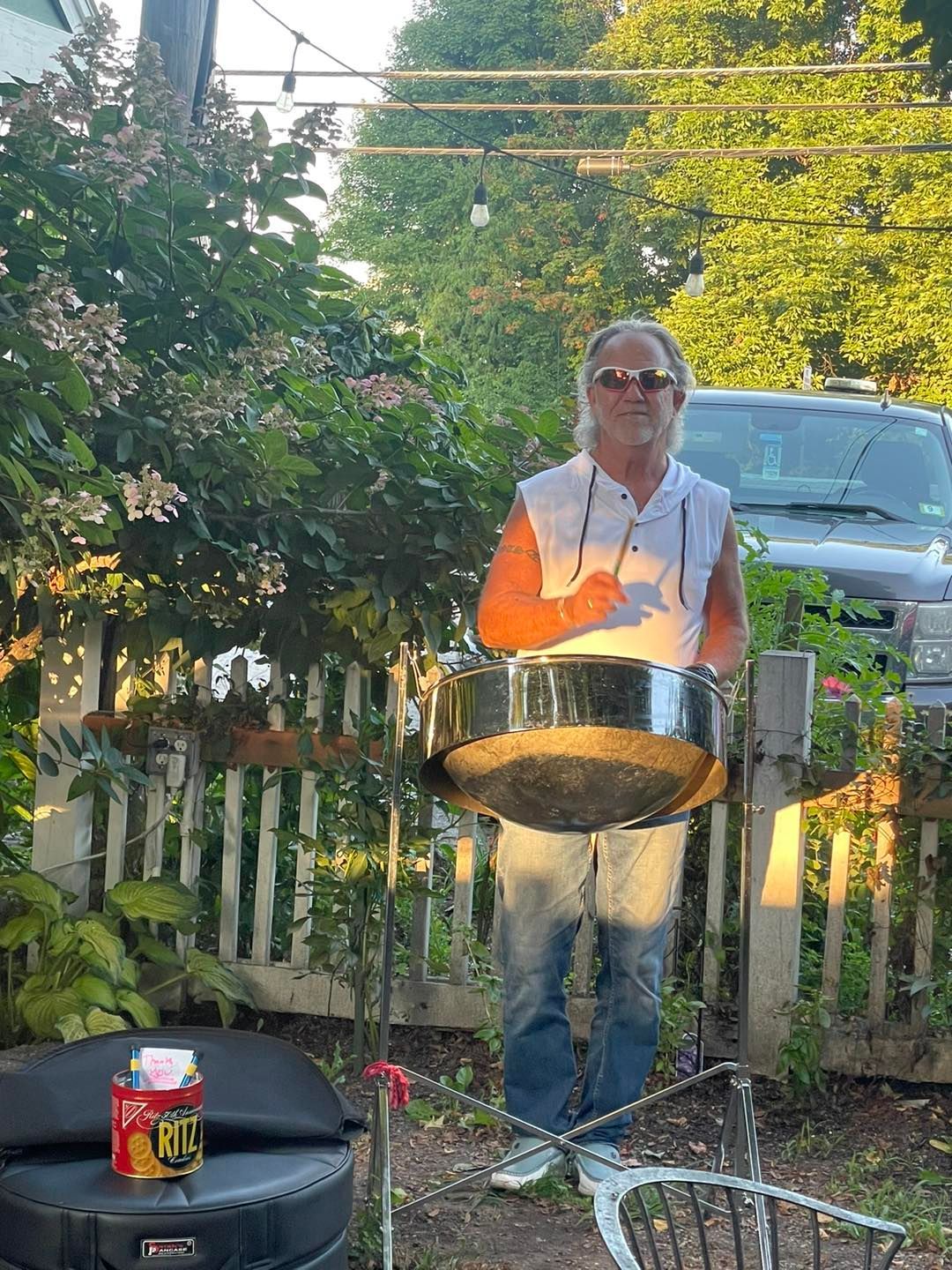A person in a sleeveless white shirt plays a steel drum outdoors in a garden setting near a white picket fence.