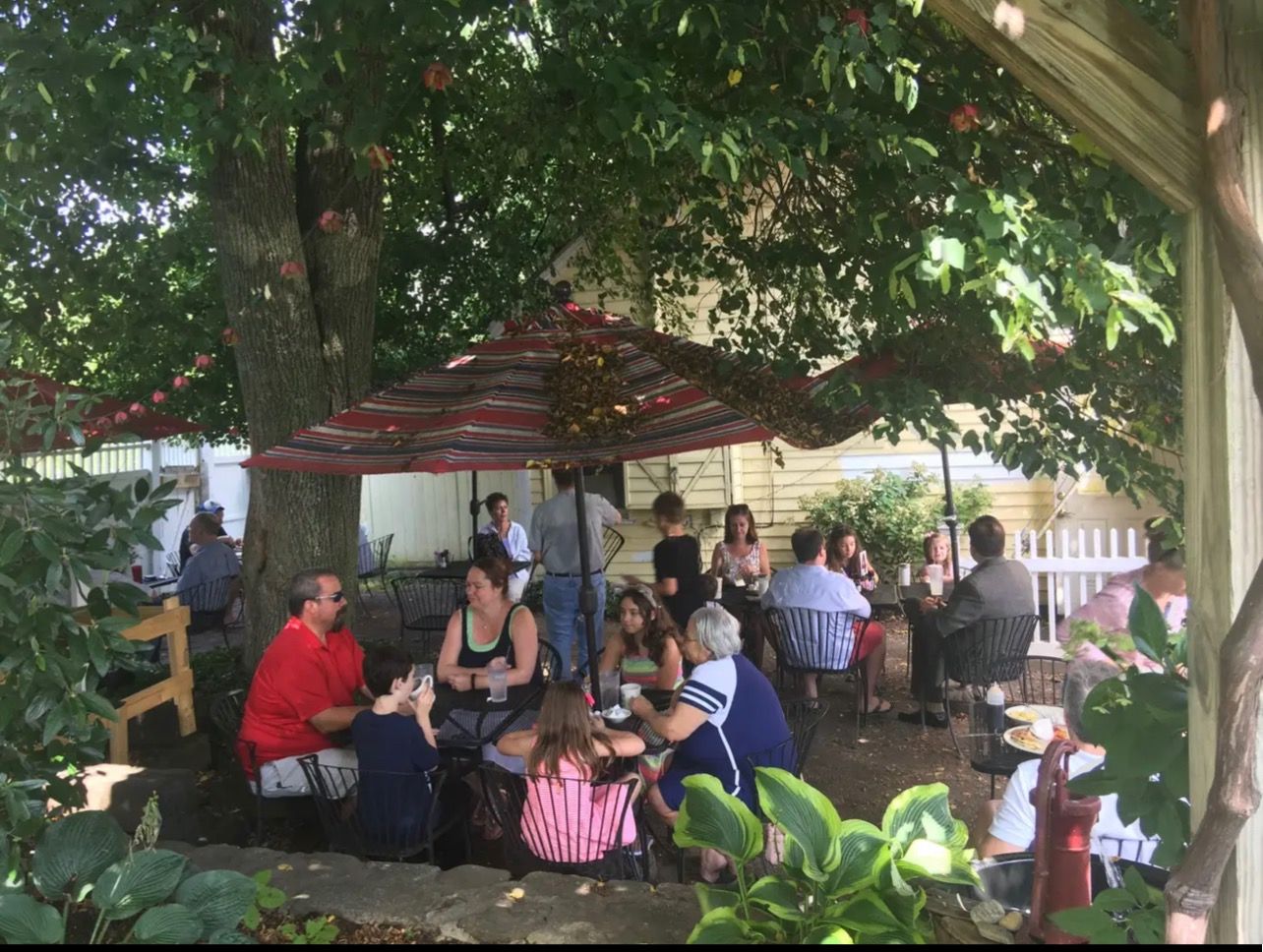 A group of people sitting at outdoor tables under a red patio umbrella and trees during a sunny day.