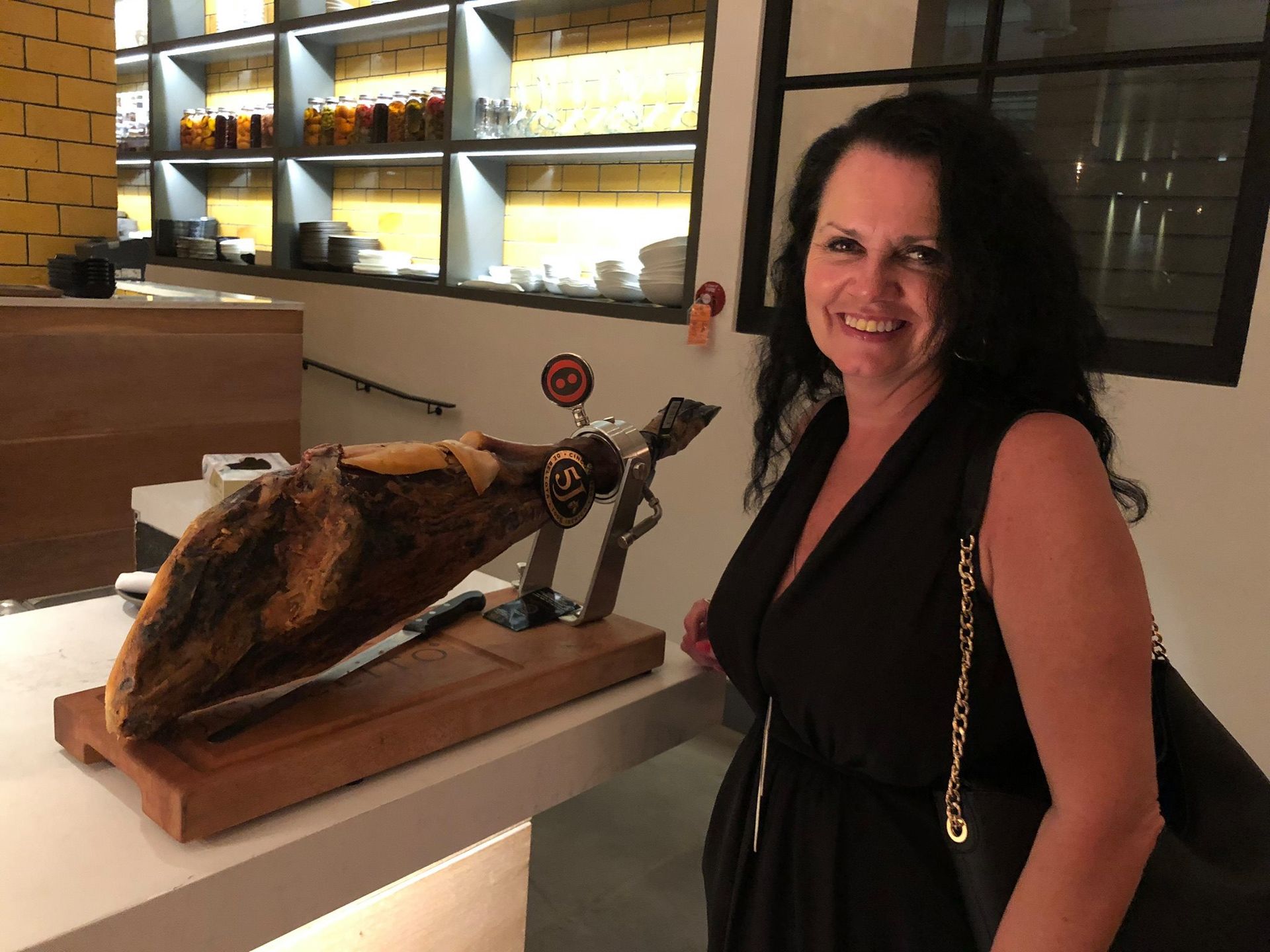 A smiling person standing in a restaurant next to a cured ham leg mounted on a wooden stand.