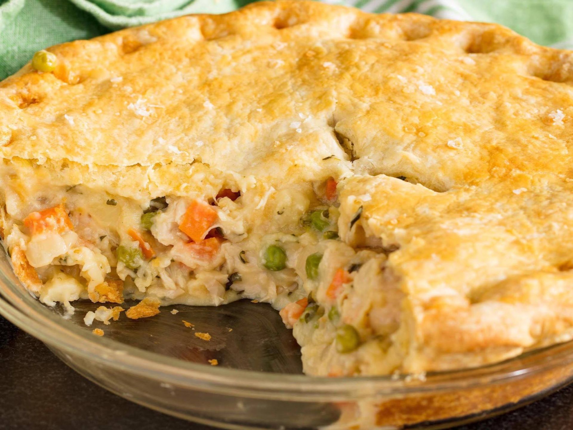 A golden-brown chicken pot pie in a glass dish with a slice cut out, showing a creamy filling with vegetables.