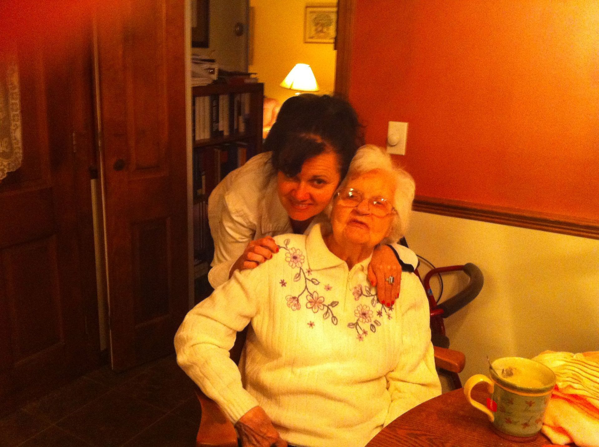 Two people embrace in a warm indoor setting with orange walls, a floral sweater, and a mug on a table in the foreground.