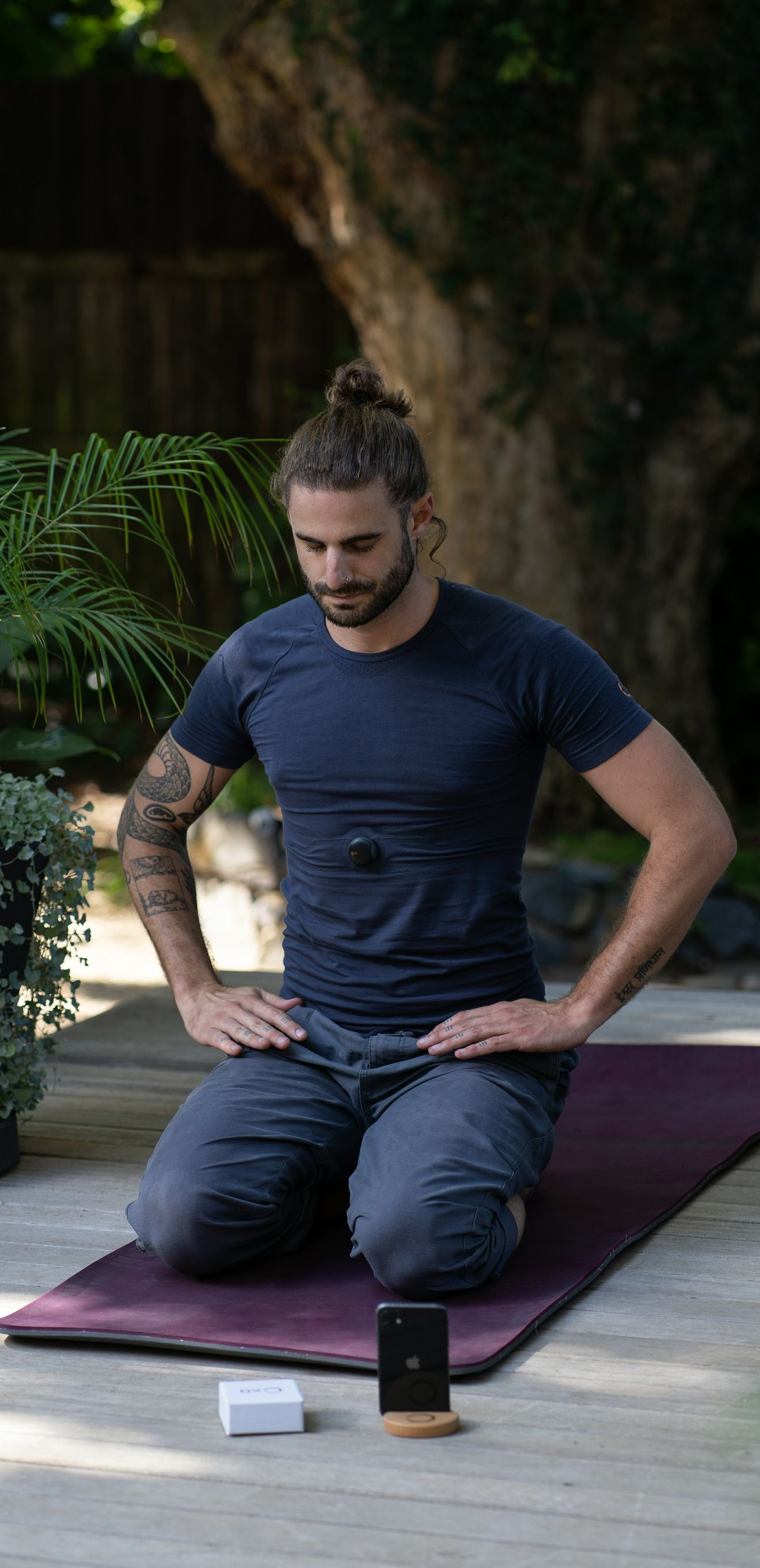 Man kneeling on a yoga mat outdoors with hands on knees, meditating. Plants and a small speaker are in view.