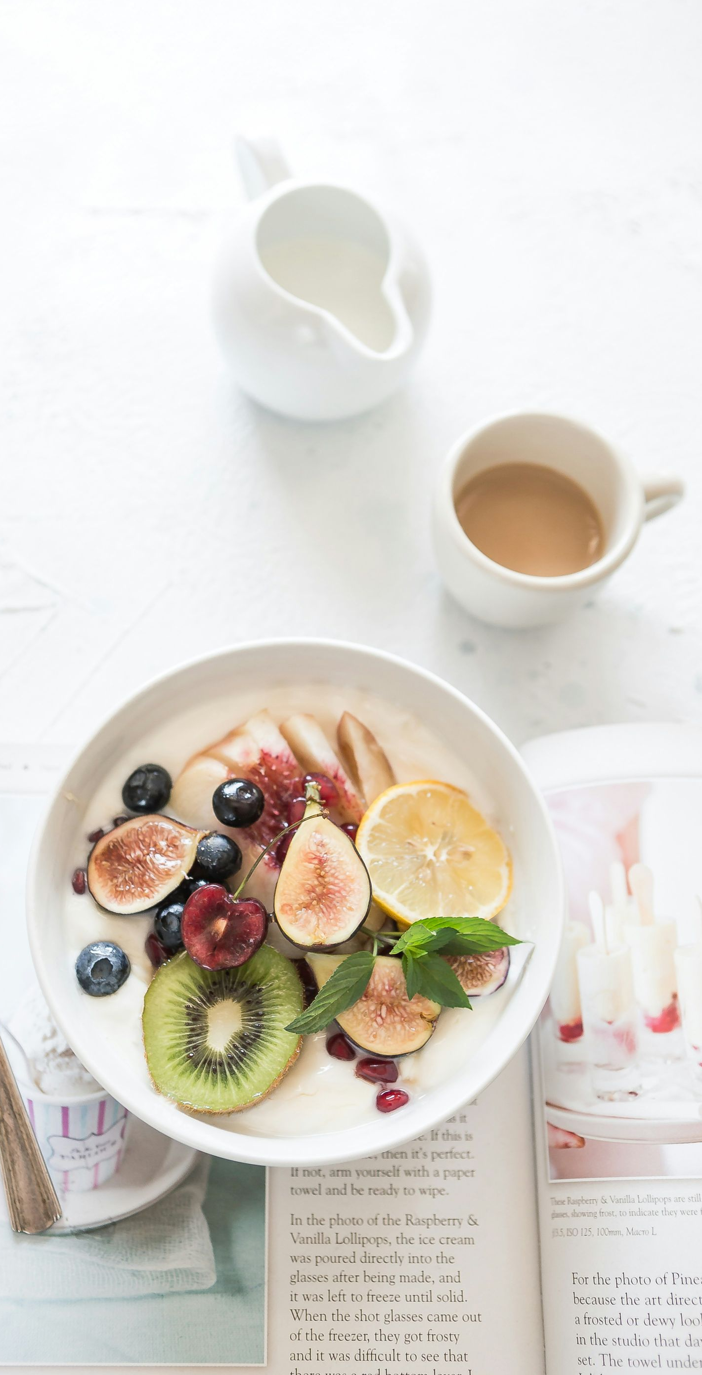 Yogurt bowl with fruit, including figs, kiwi, and berries, with coffee and a pitcher on a white surface.