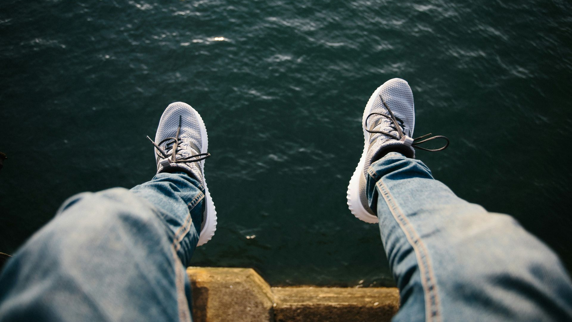 Person's legs in blue jeans and white sneakers hanging over water.
