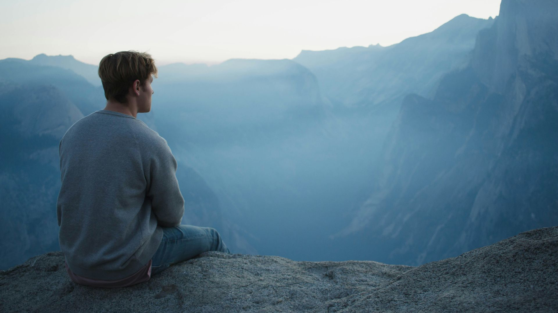 Person seated on a cliff overlooking mountains and a misty valley.
