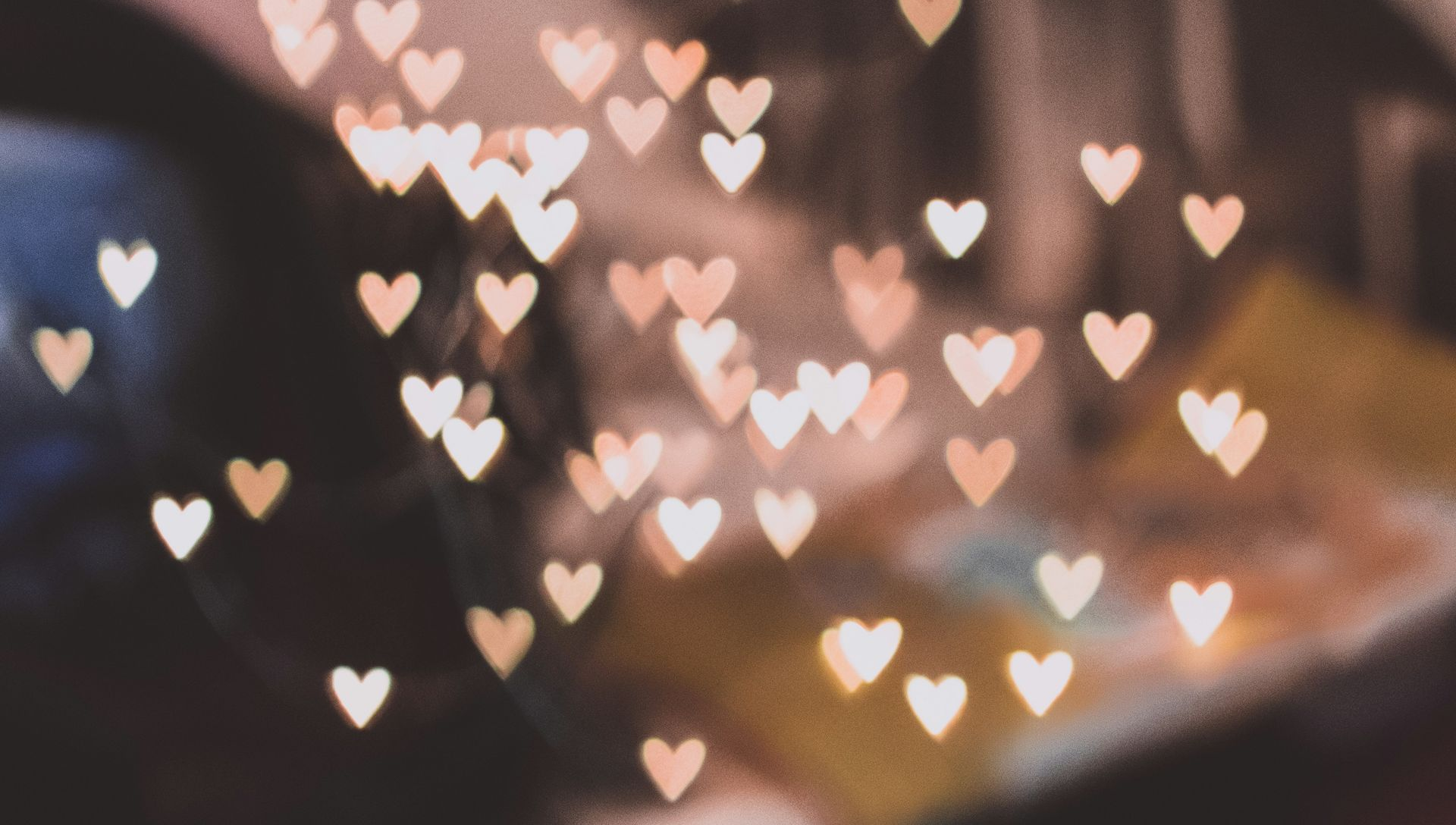 Blurred shot of glowing heart-shaped lights, soft peach and white hues. Background includes blurry objects, presumably an indoor setting.