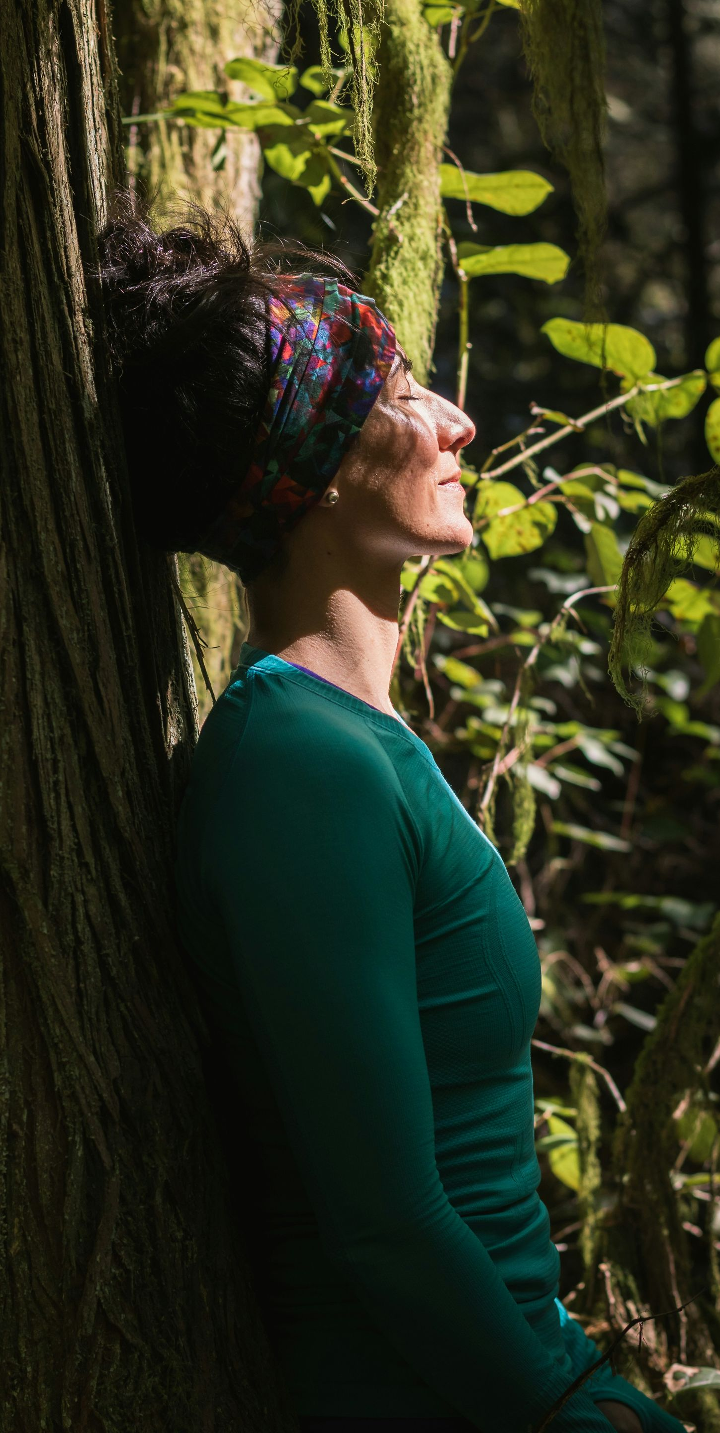 Woman leaning against tree, eyes closed, sunlight, forest, wearing teal top, patterned head wrap.