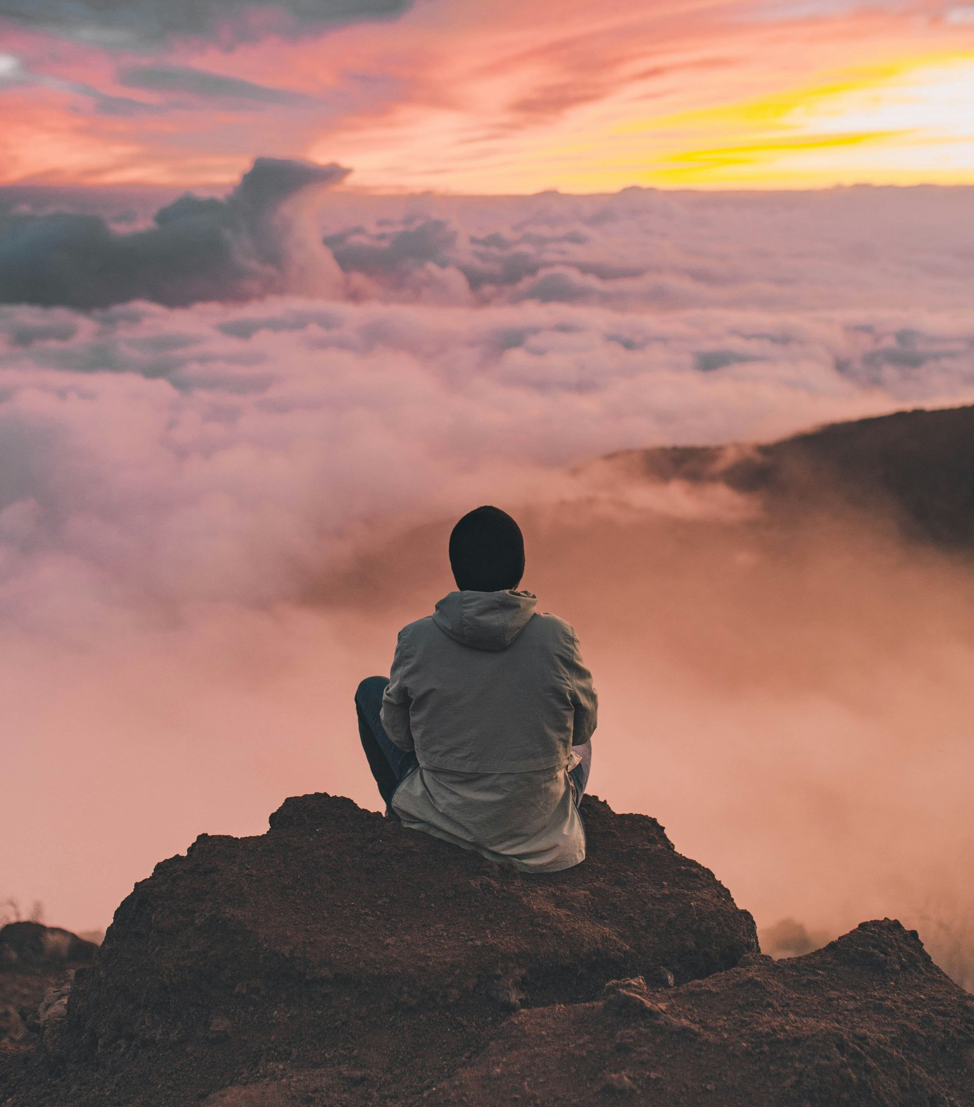 Person seated on a rock, back to the camera, overlooking clouds at sunset.