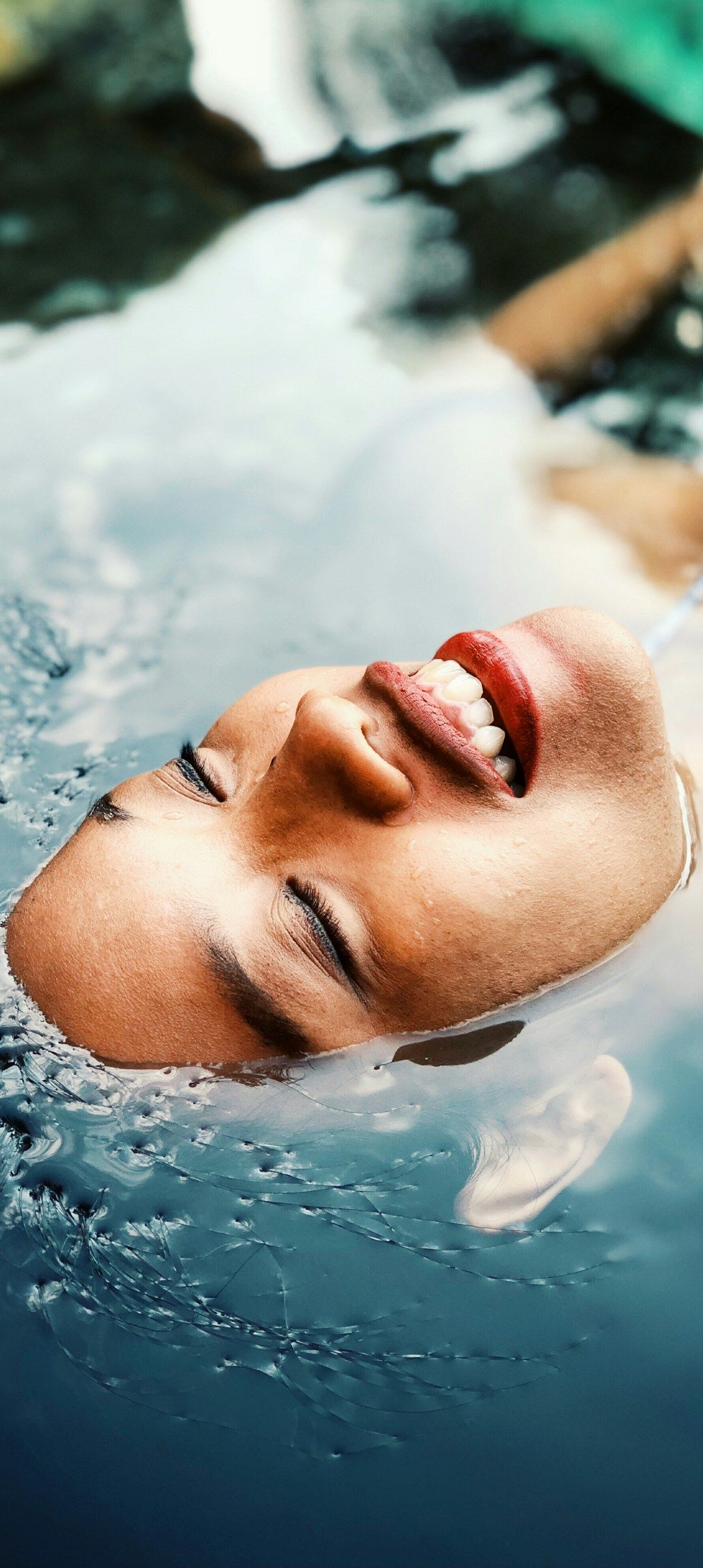 A person floats in water, face visible, mouth open. Soft lighting, blue water, blurred background.