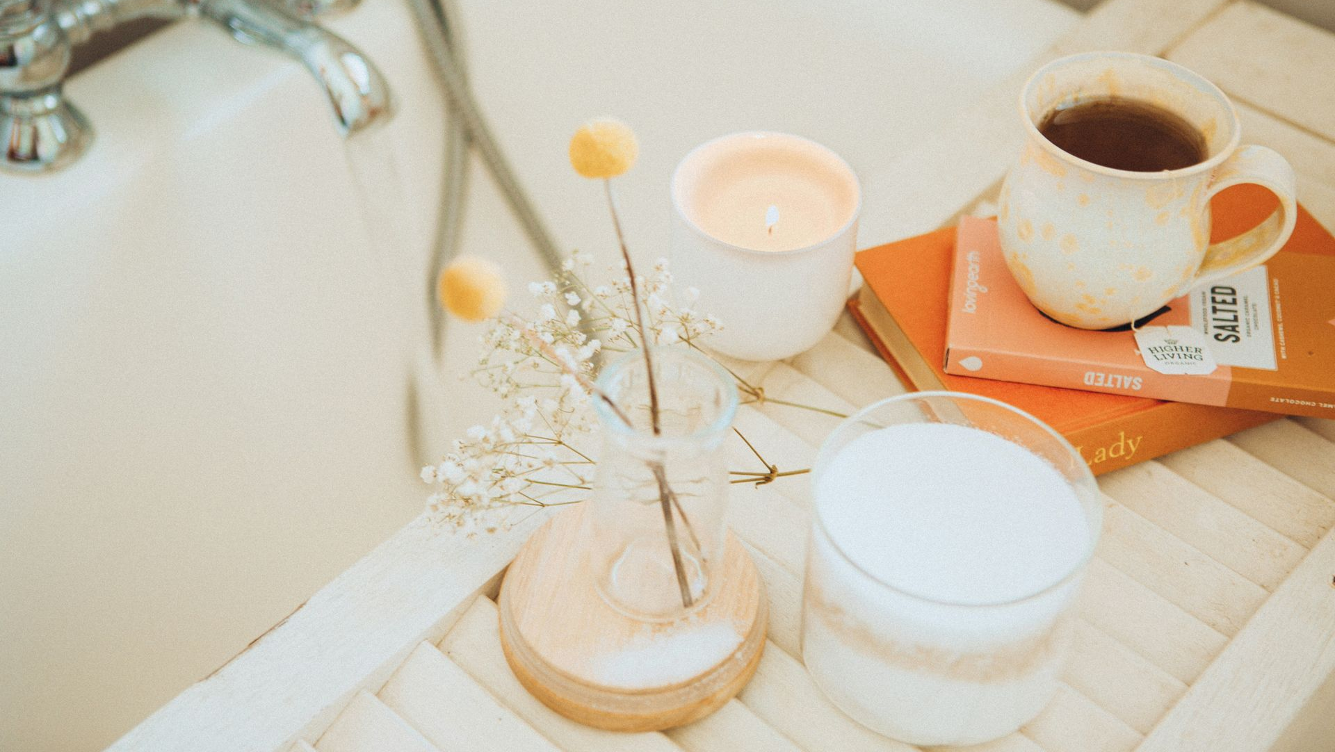 Bathtub tray with a candle, flowers, a mug of tea, books, and a glass of bath salts.