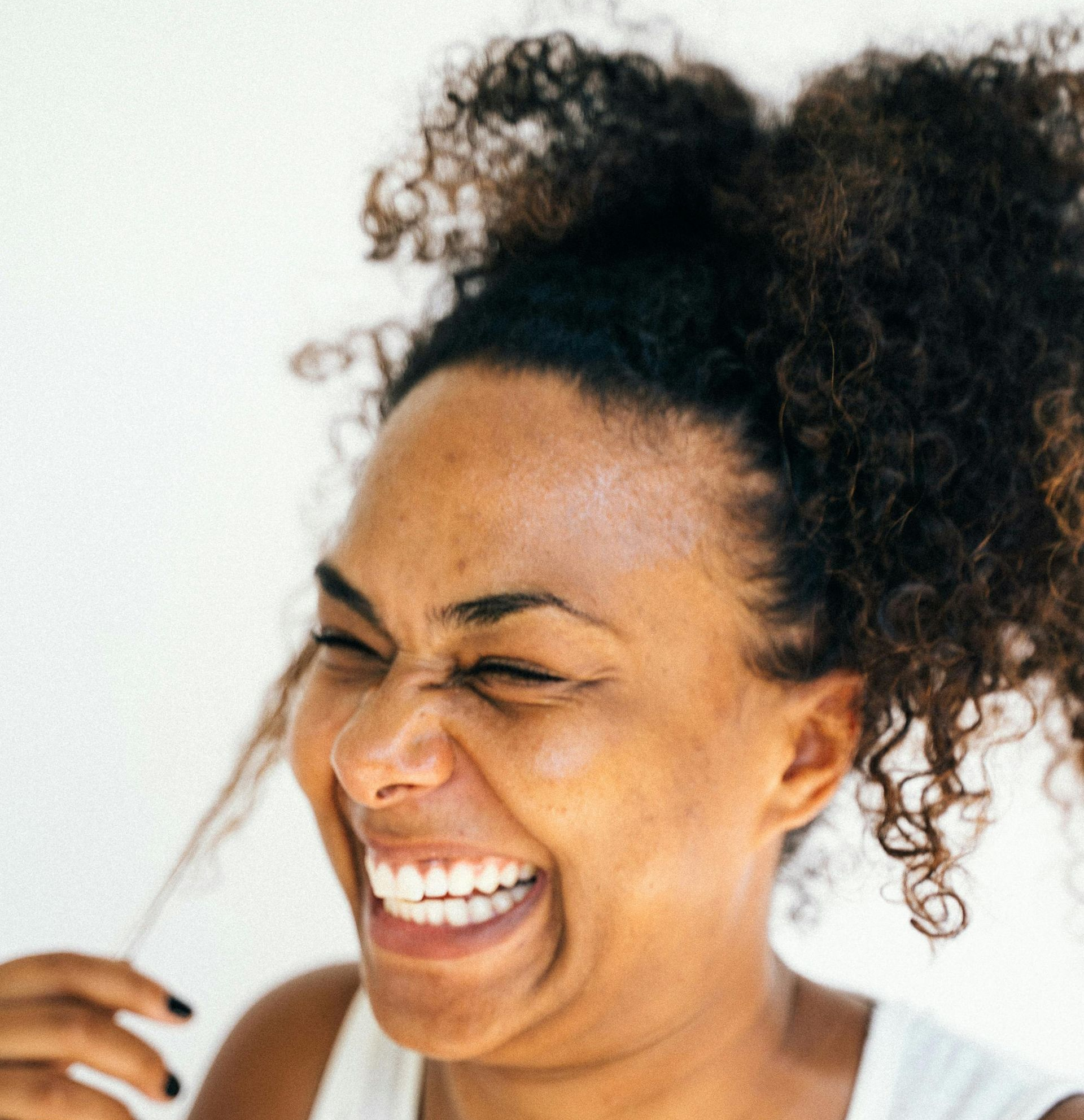 Woman with curly dark hair laughing, eyes closed, white teeth visible.