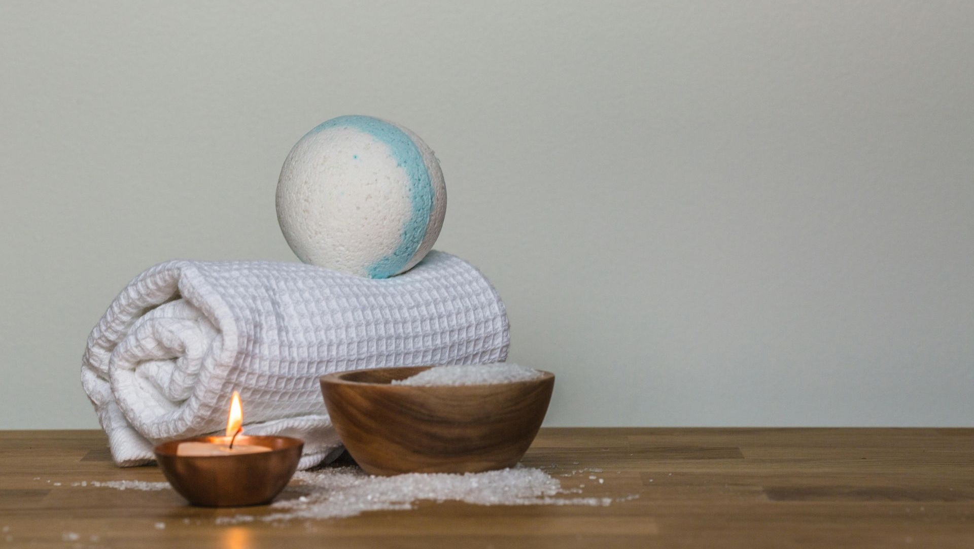 Rolled white towel, bath bomb, wooden bowls with salt and candle, on wood surface.