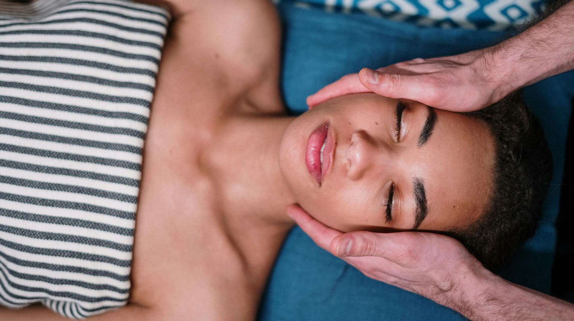 Person receiving a facial massage, hands on face, eyes closed, resting on blue pillow, wrapped in towel.