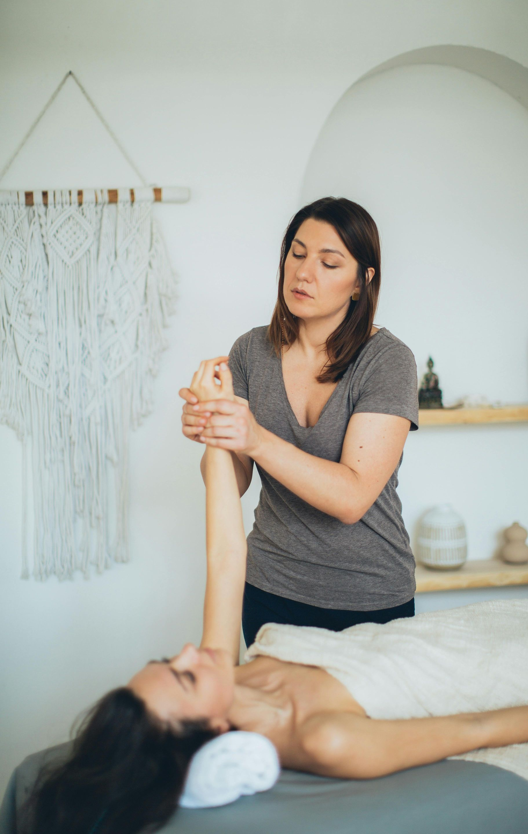 Woman giving arm massage to another woman on a massage table. White-walled room with macrame.