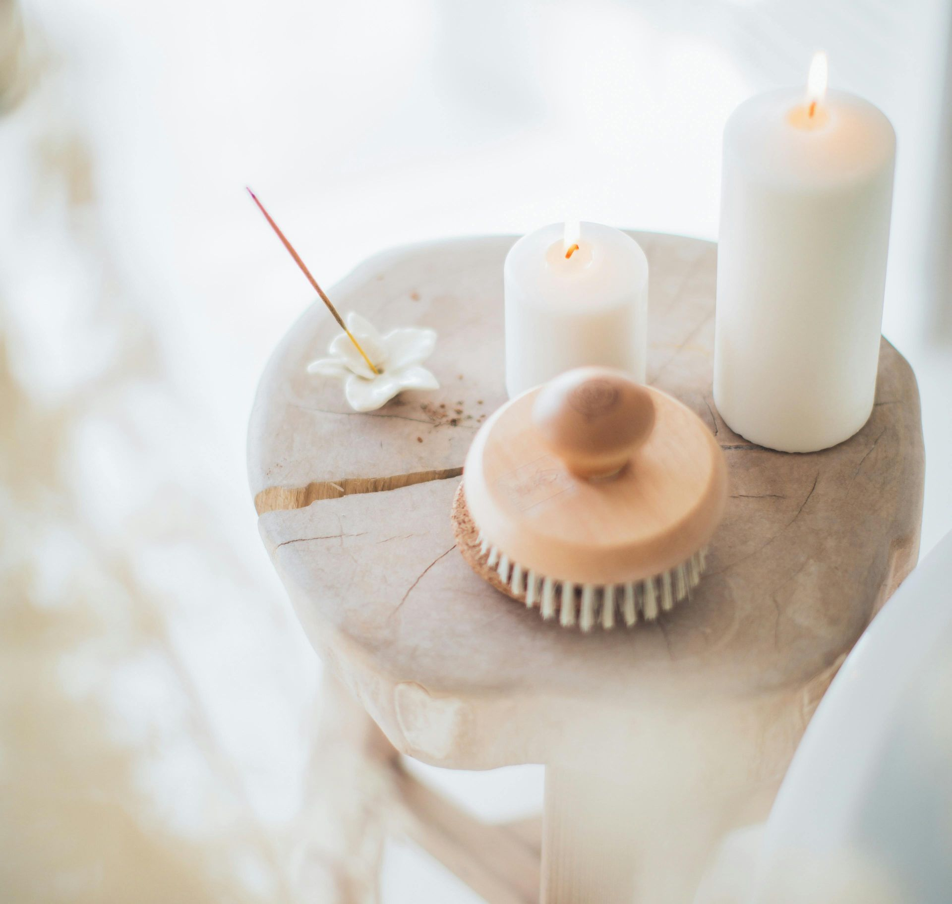 White candles, a brush, incense, and a flower on a small, round, wooden table in a well-lit space.