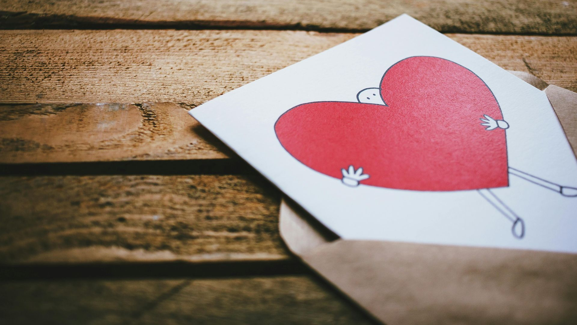 Card with stick figure hugging red heart, in a brown envelope on a wood surface.