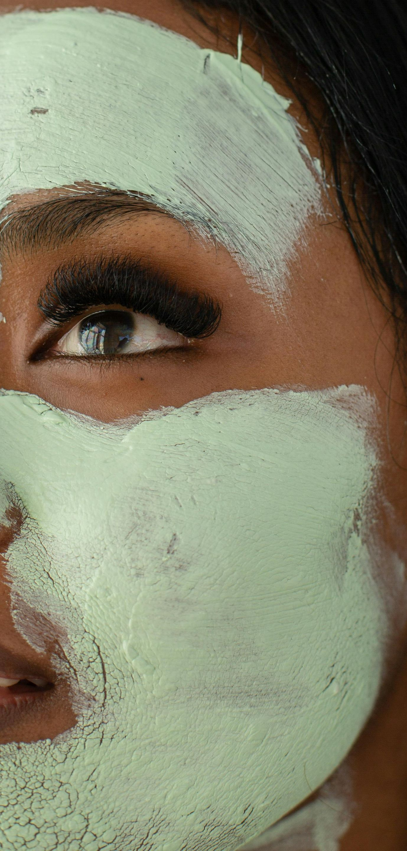 Woman's face with light green facial mask applied. She smiles with eye makeup.