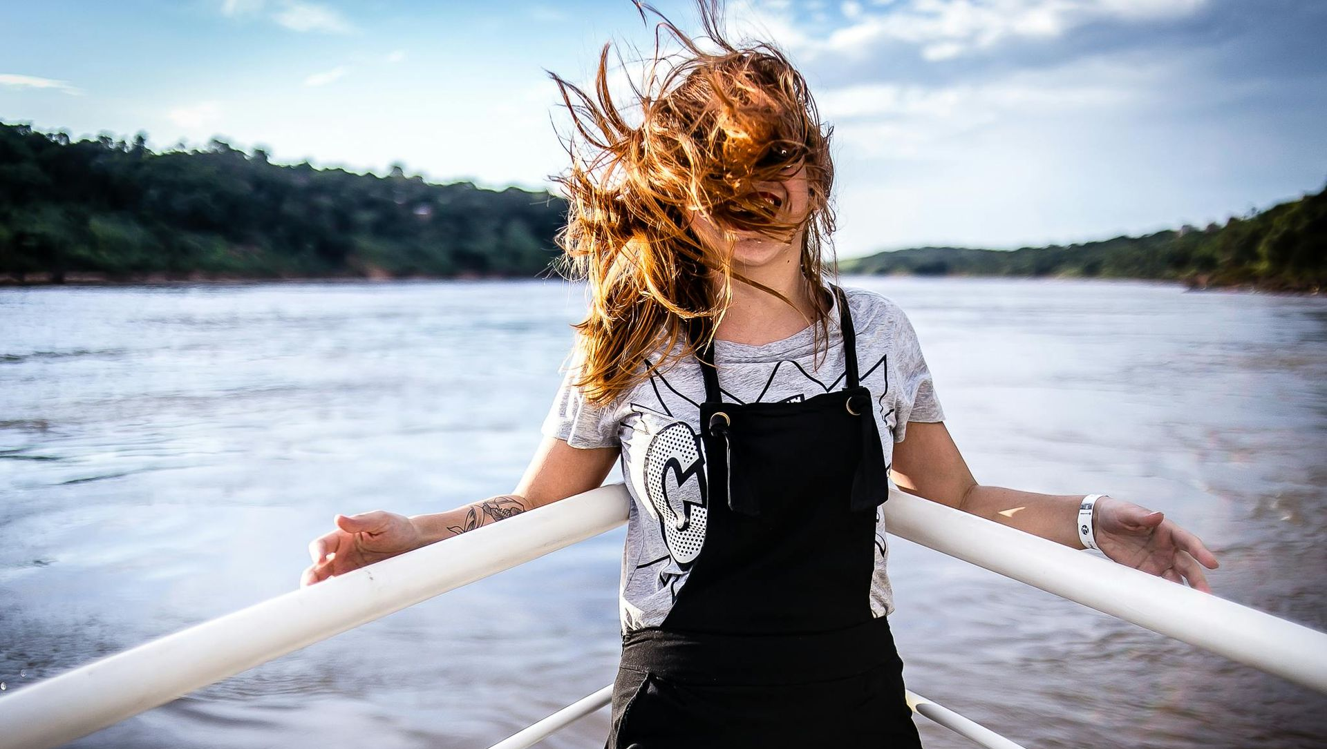 Woman on a boat with windblown hair, smiling, arms outstretched. White railing, river, and trees in background.
