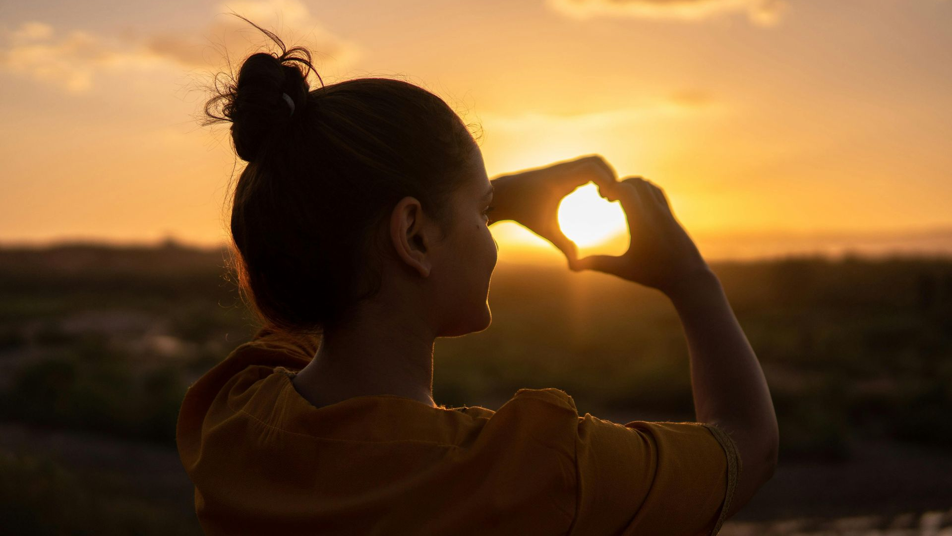 Woman holding hands in a heart shape framing the sun during a golden sunset over a landscape.