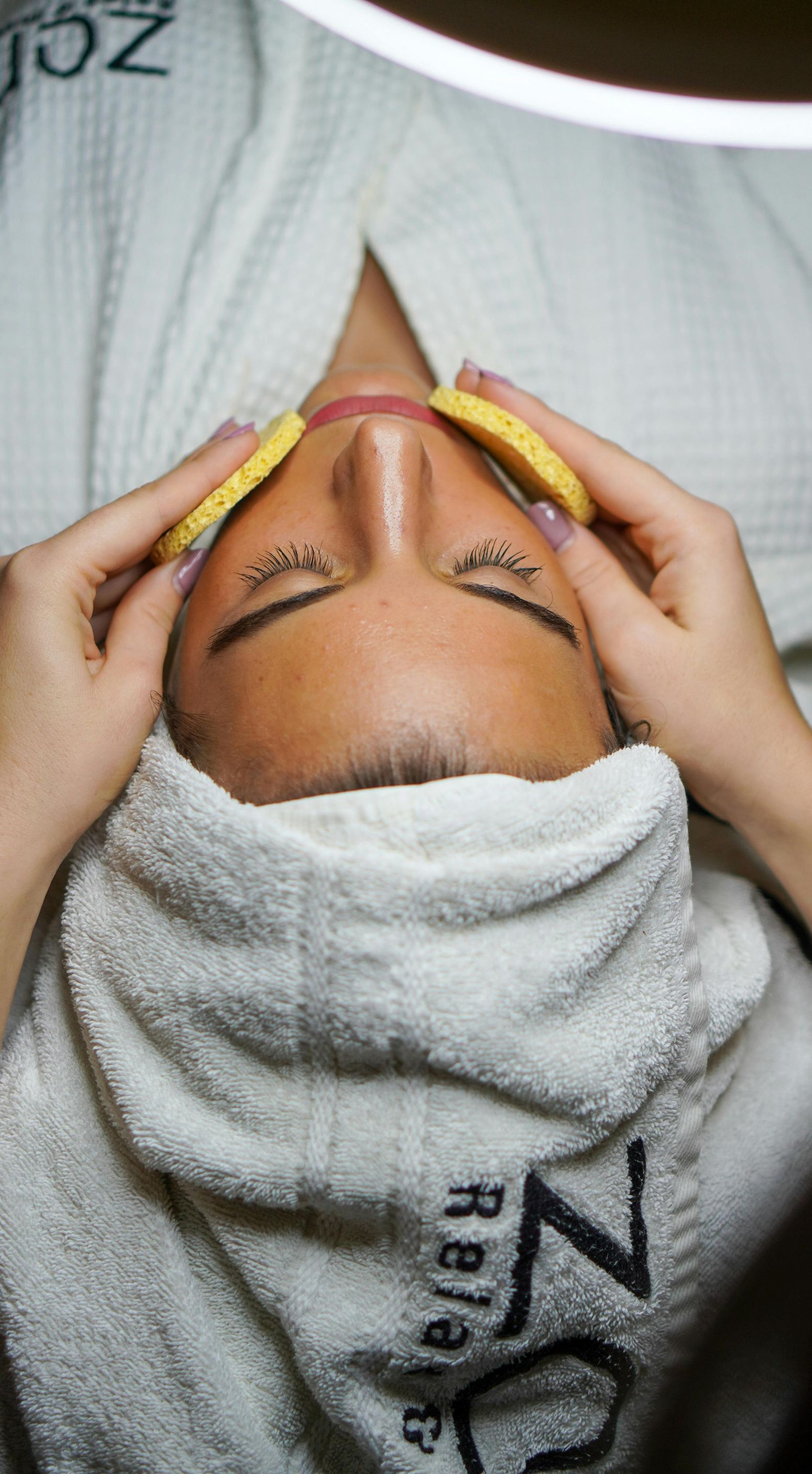 Person receiving facial treatment, covered in towel, sponges on face. Spa setting, overhead light.