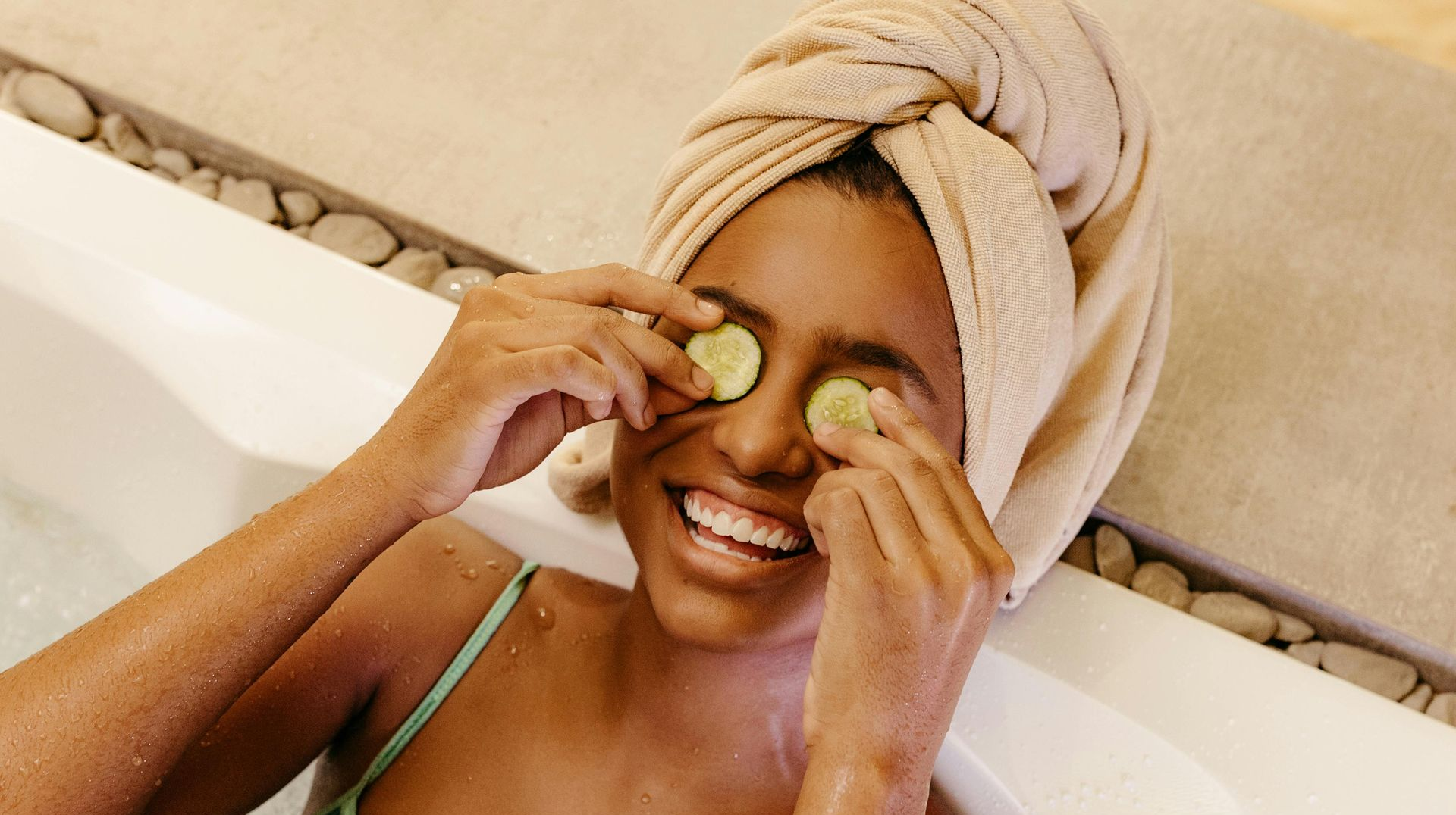 Woman in a bathtub, towel on head, holding cucumber slices over her eyes, smiling.